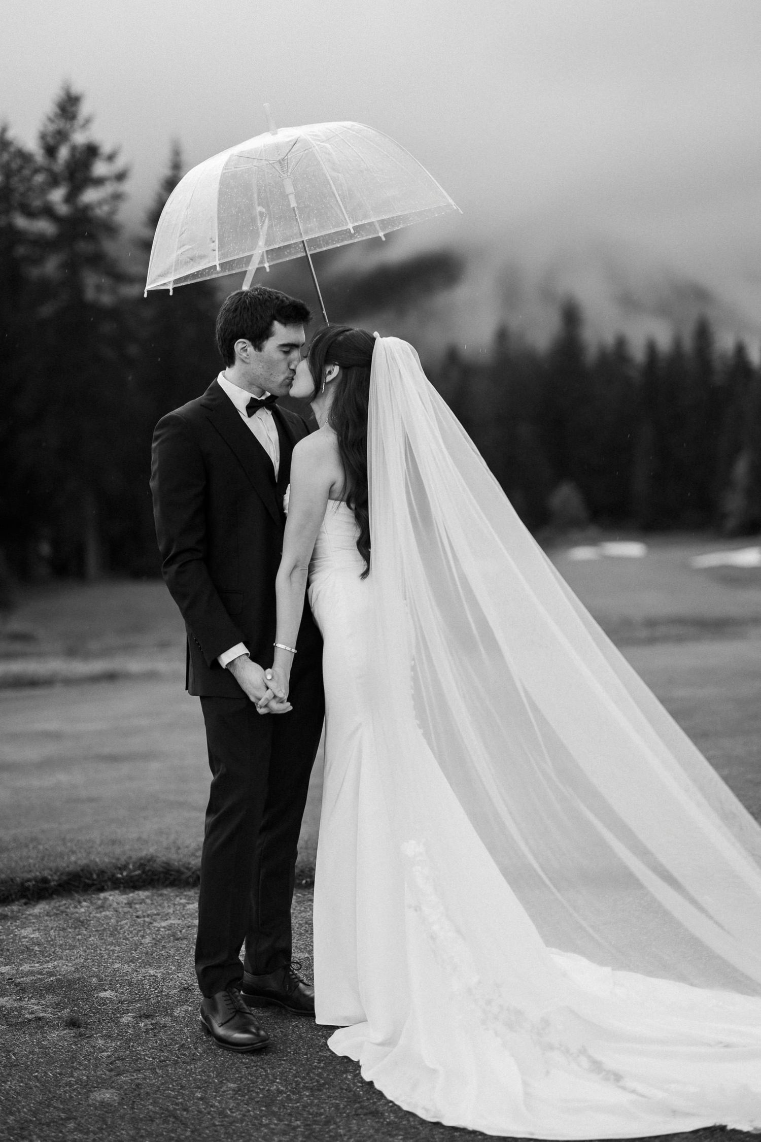 Black and white photo of bride and groom kissing under clear umbrella on rainy wedding day with misty mountain backdrop.