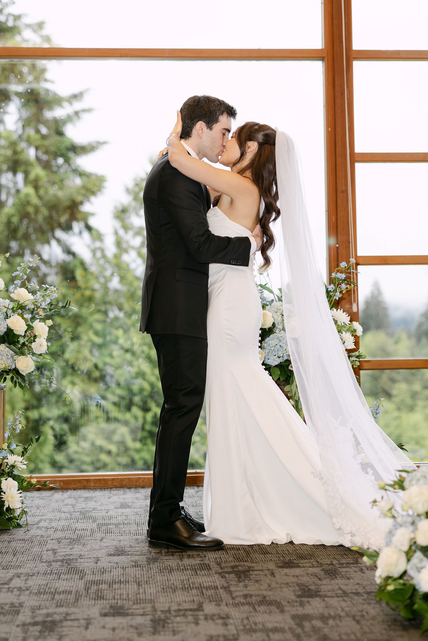 Bride and groom share first kiss at altar with floral arrangements and forest view window backdrop.