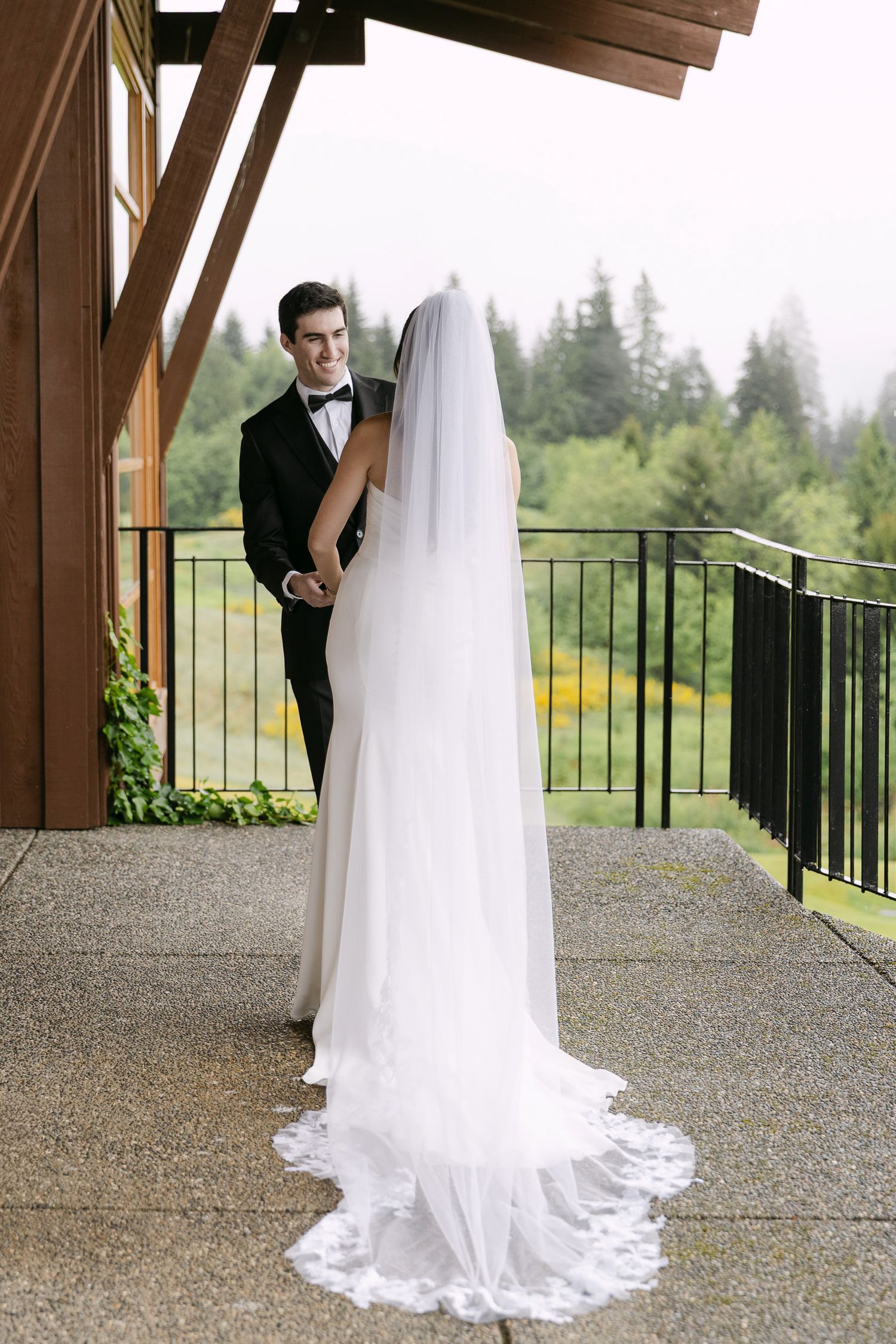 Bride in white gown with long lace veil holds groom in tuxedo on outdoor deck surrounded by lush green trees.