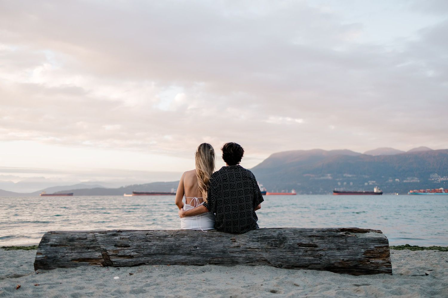 Two people sit together on a rocky beach overlooking the ocean at sunset with mountains in the distance.