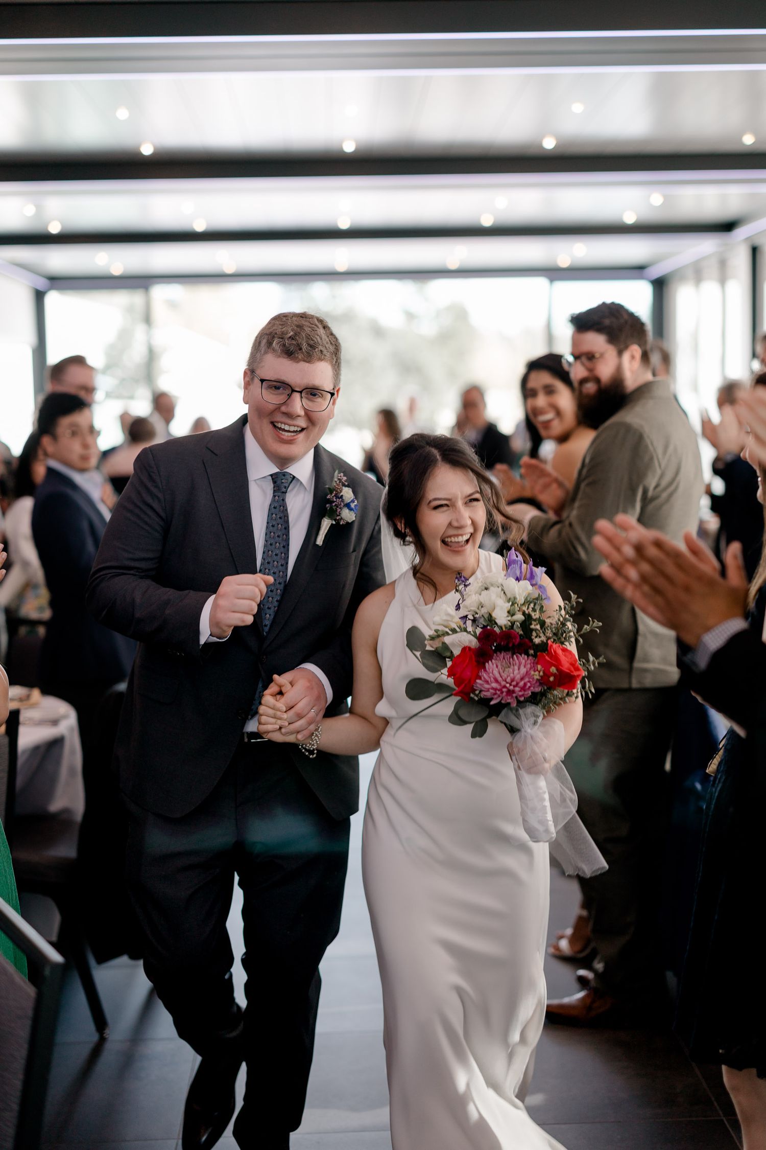 A joyful couple walks down the aisle at their wedding reception while guests clap and celebrate.