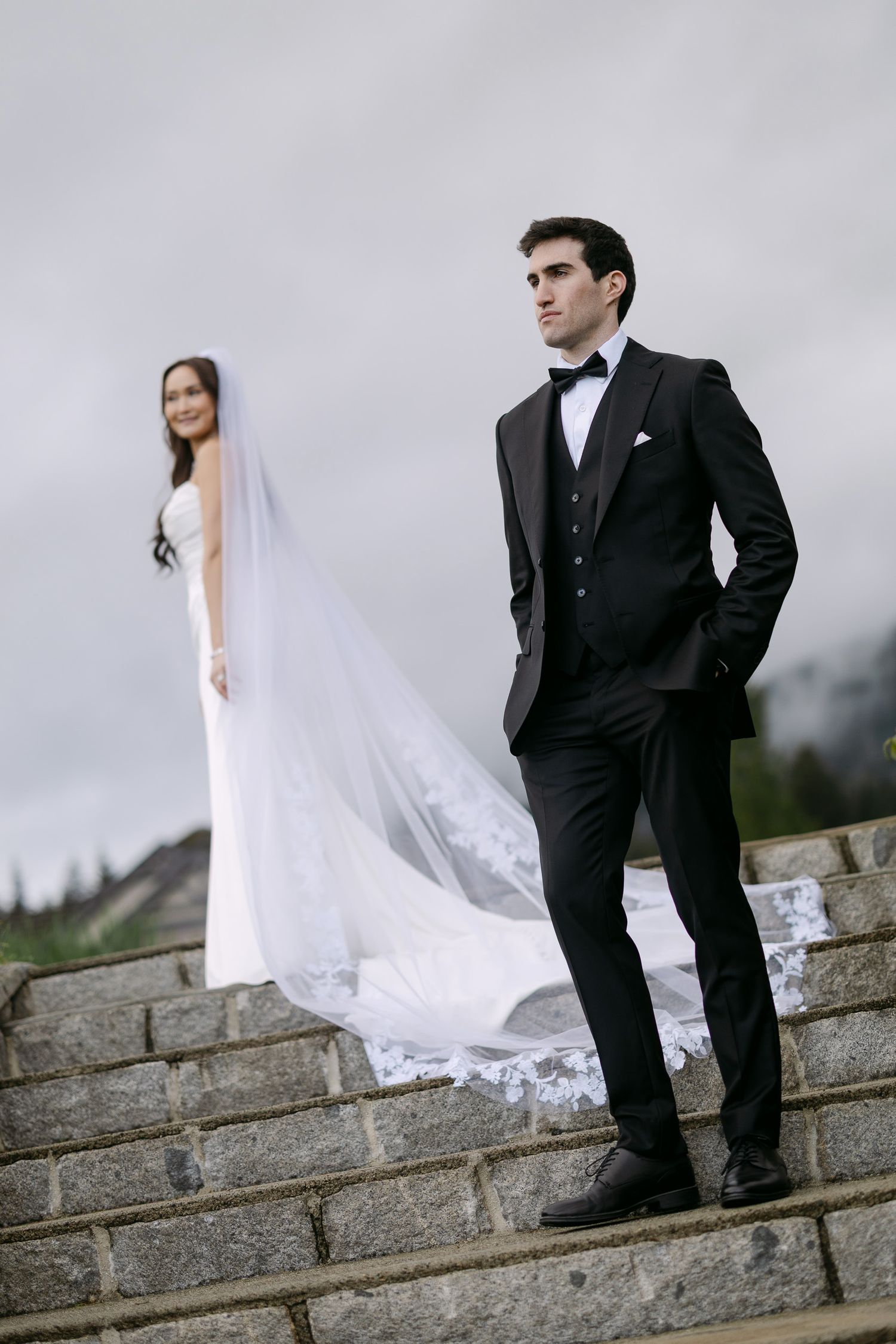 Groom in black tuxedo stands on stone steps with bride in white gown and cathedral veil behind him.
