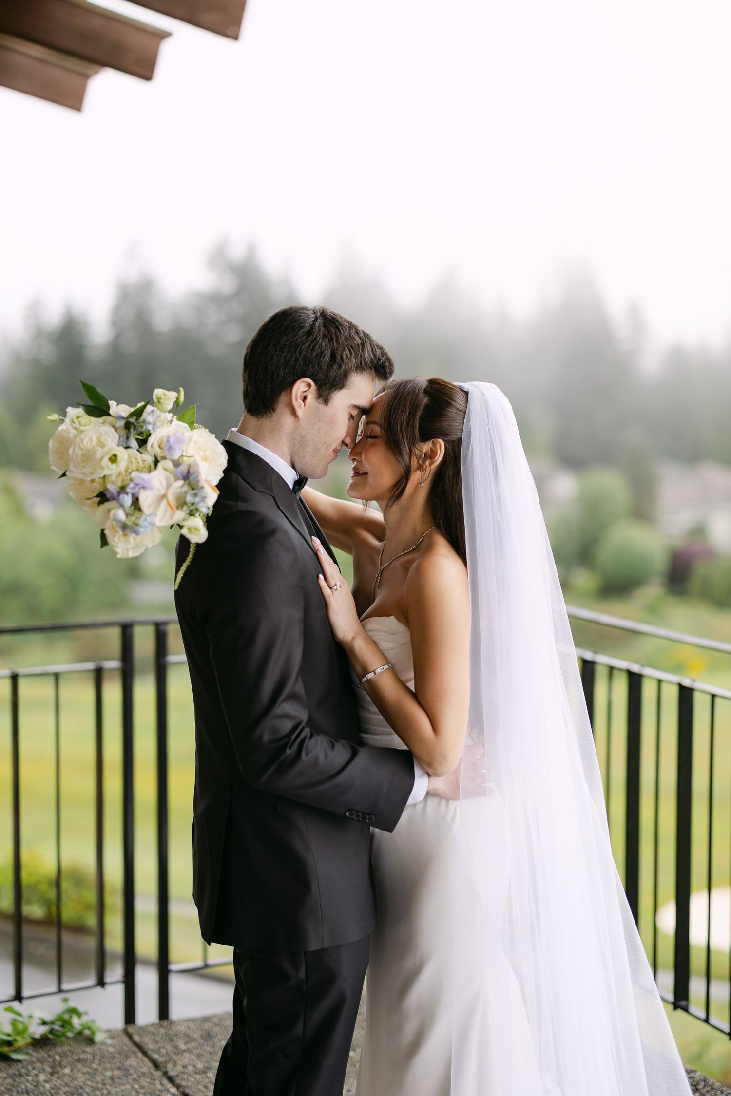 Bride and groom share a romantic moment on a balcony, with bride holding white bouquet and wearing veil.