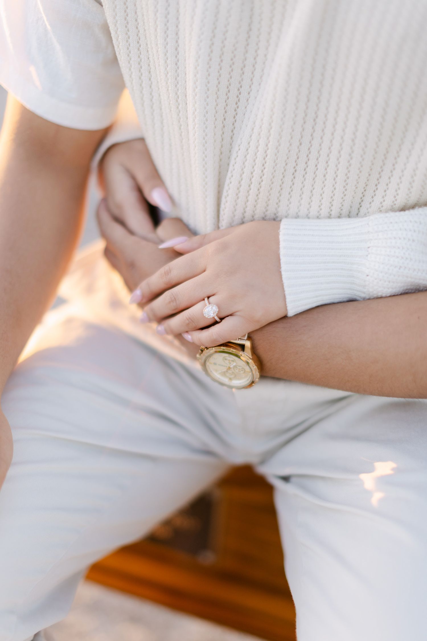 Close-up of a woman's hand with a sparkling diamond engagement ring and gold watch resting on white linen pants.