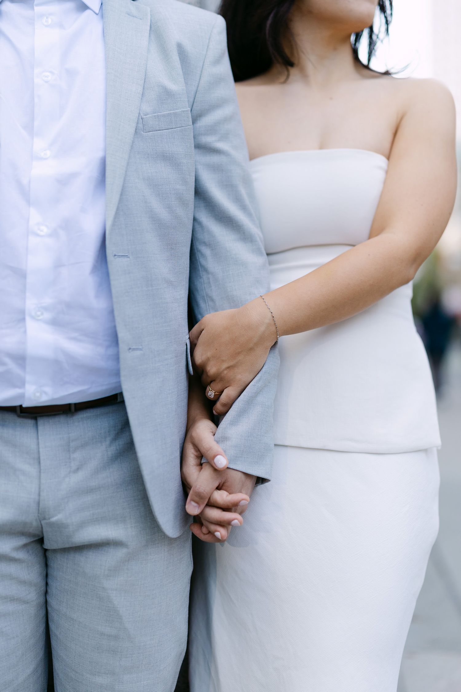 Close-up of a couple holding hands, she in a white strapless dress and he in a light gray suit and white shirt.