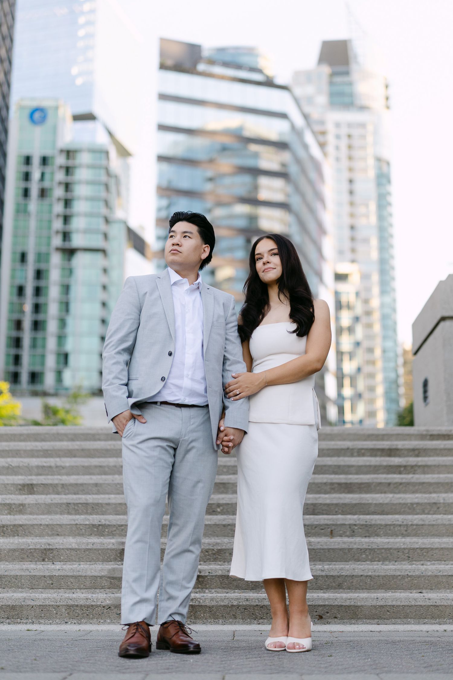 Couple holding hands on concrete steps in a gray suit and white dress, with modern glass skyscrapers rising behind them.
