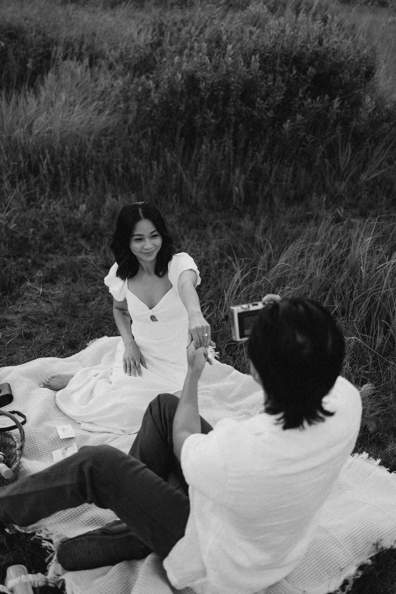 Black and white photo of a couple sharing a quiet moment on a picnic blanket in a grassy field while he holds a camera.