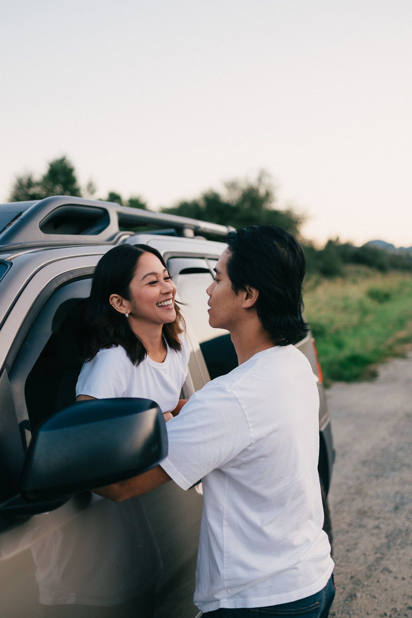 A smiling woman sits in a car window as her partner leans in close beside a rural roadside at sunset.