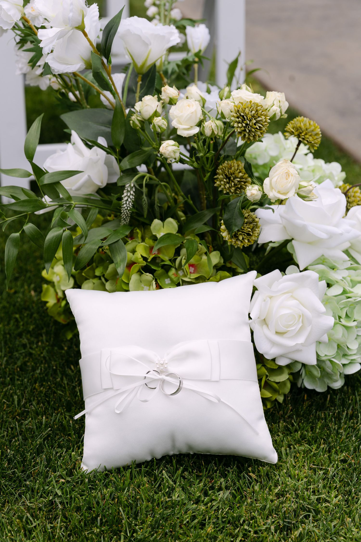 White satin ring bearer pillow with wedding rings rests against lush floral arrangement on green grass.