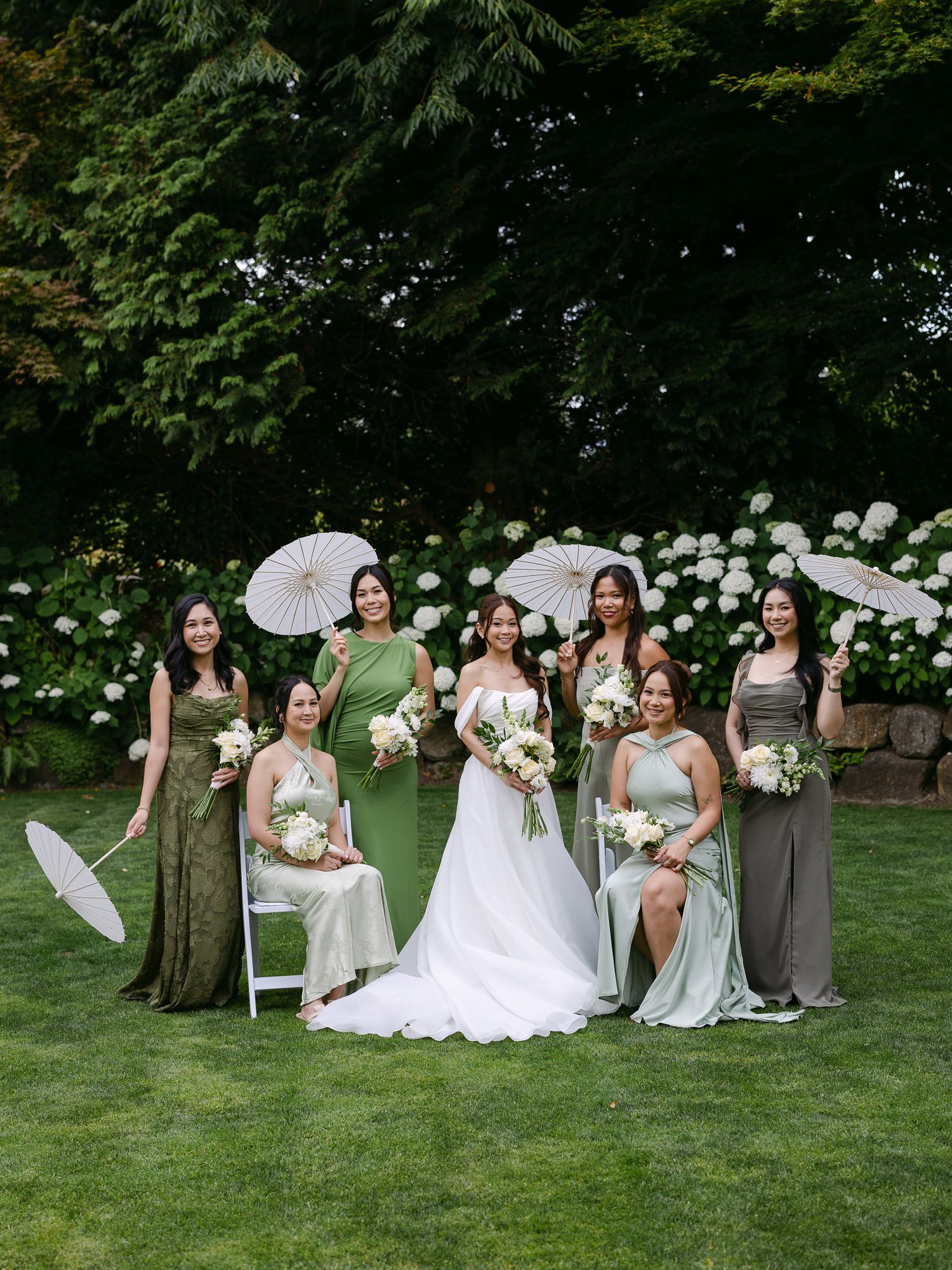 Bride in white gown with bridesmaids in green dresses holding parasols and bouquets on a lawn with hydrangeas.