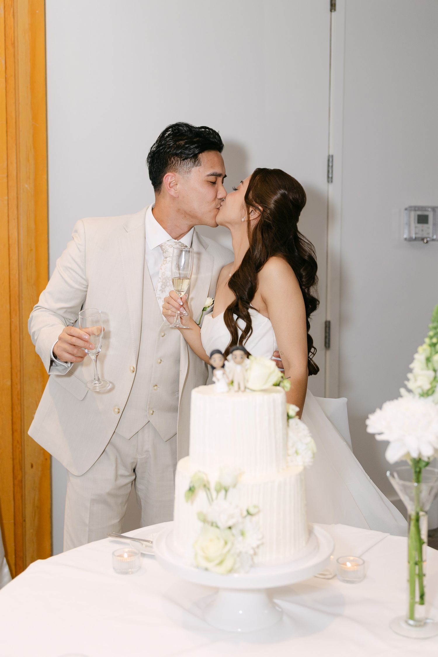 Bride and groom kissing behind a white two-tier wedding cake while holding champagne glasses at their reception.