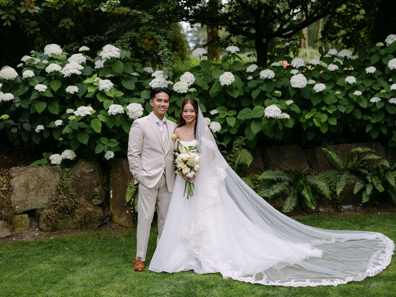 Bride and groom pose together in a lush garden with white hydrangeas, bride wearing a flowing gown with cathedral veil.