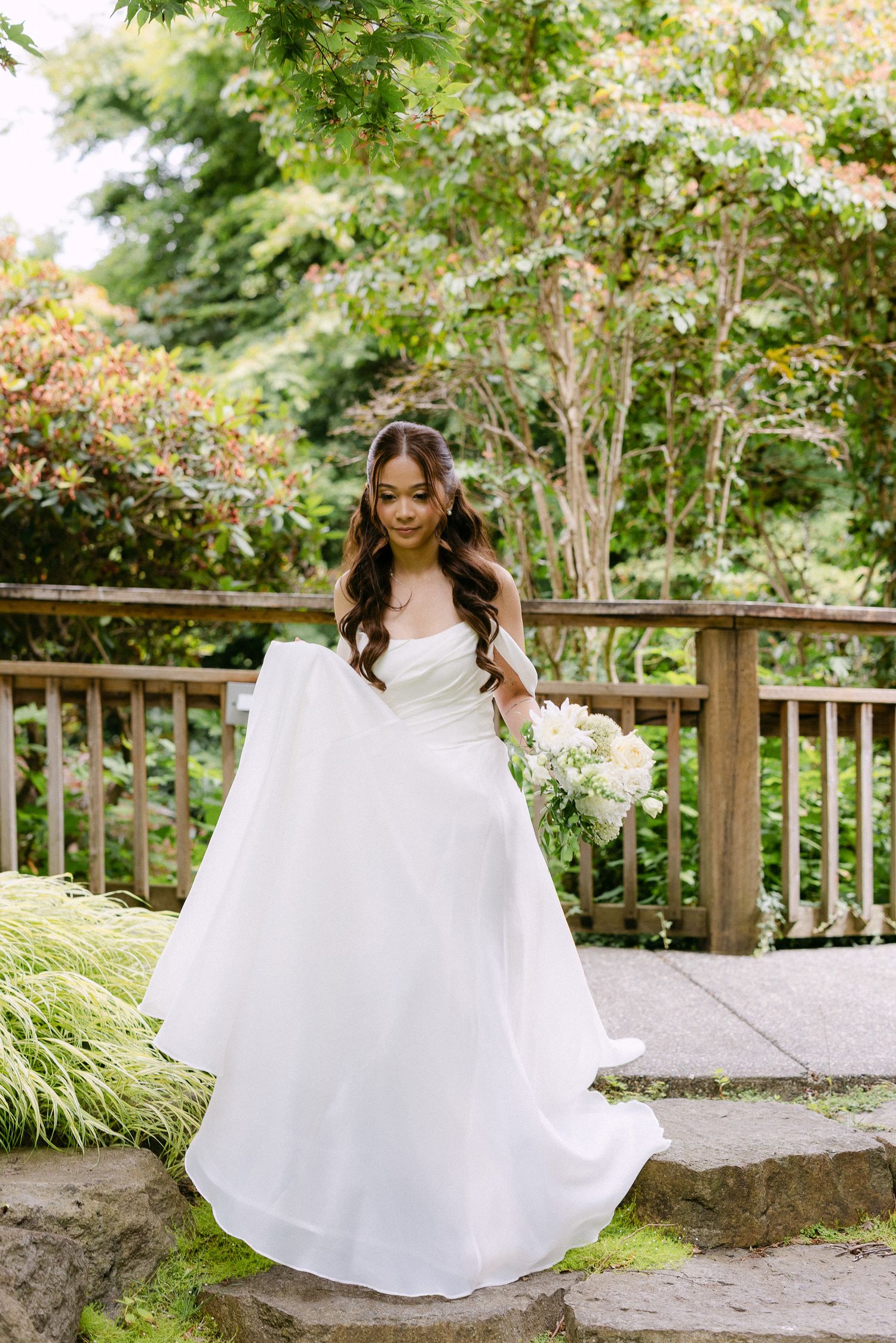Bride in white strapless ball gown holding bouquet on stone steps in lush garden setting.