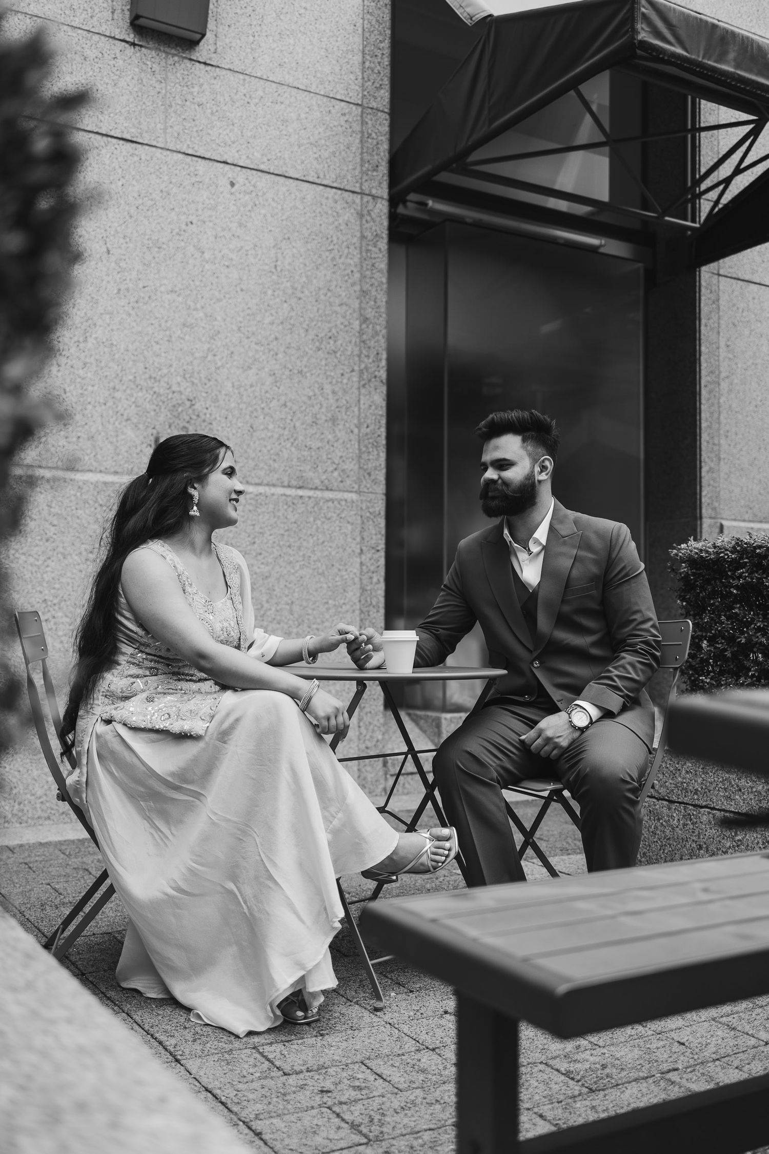 A couple sits together at an outdoor cafe table in black and white, sharing an intimate conversation.
