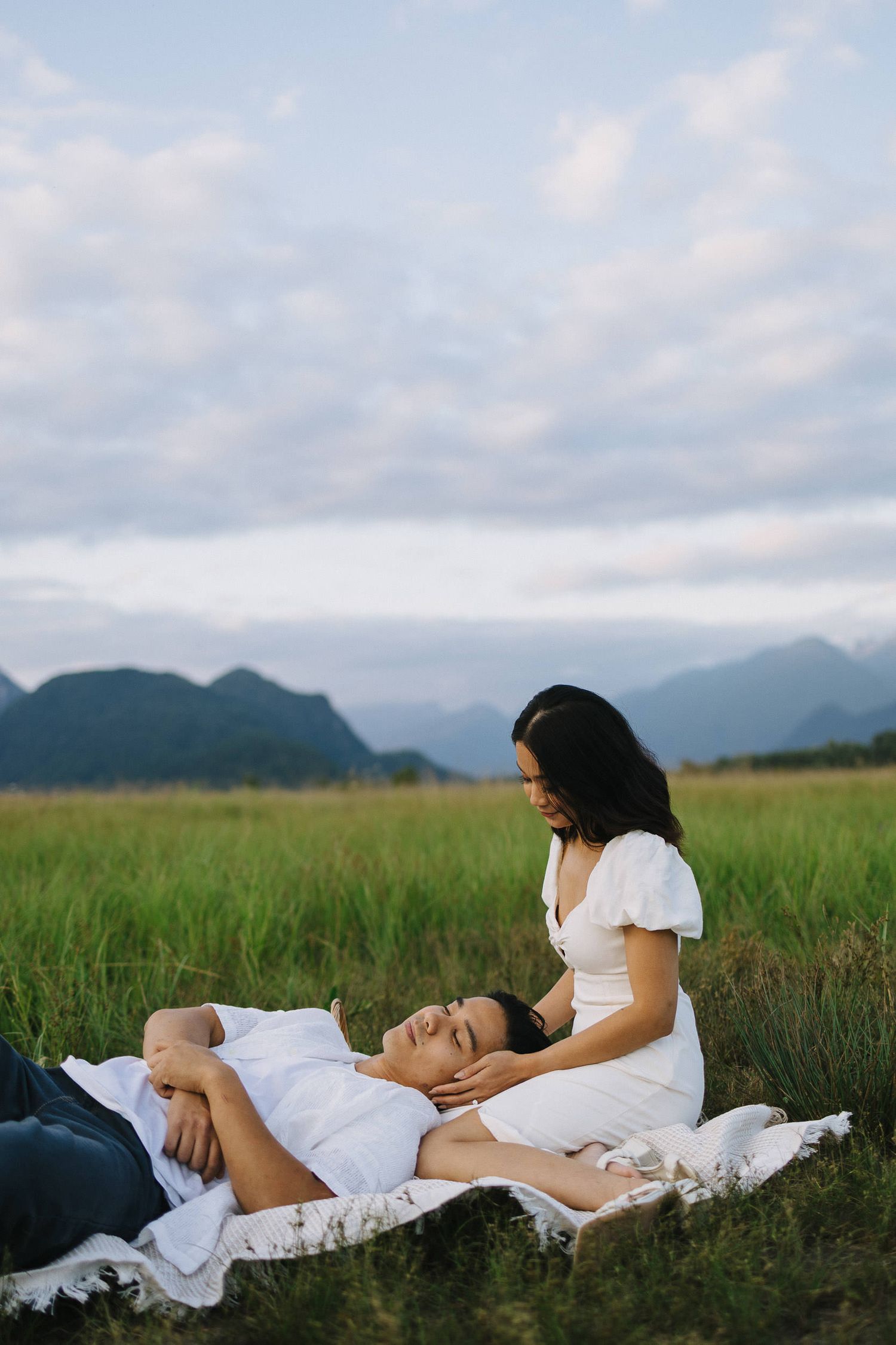 A couple in white clothing lies together in a peaceful green meadow with mountains in the background.