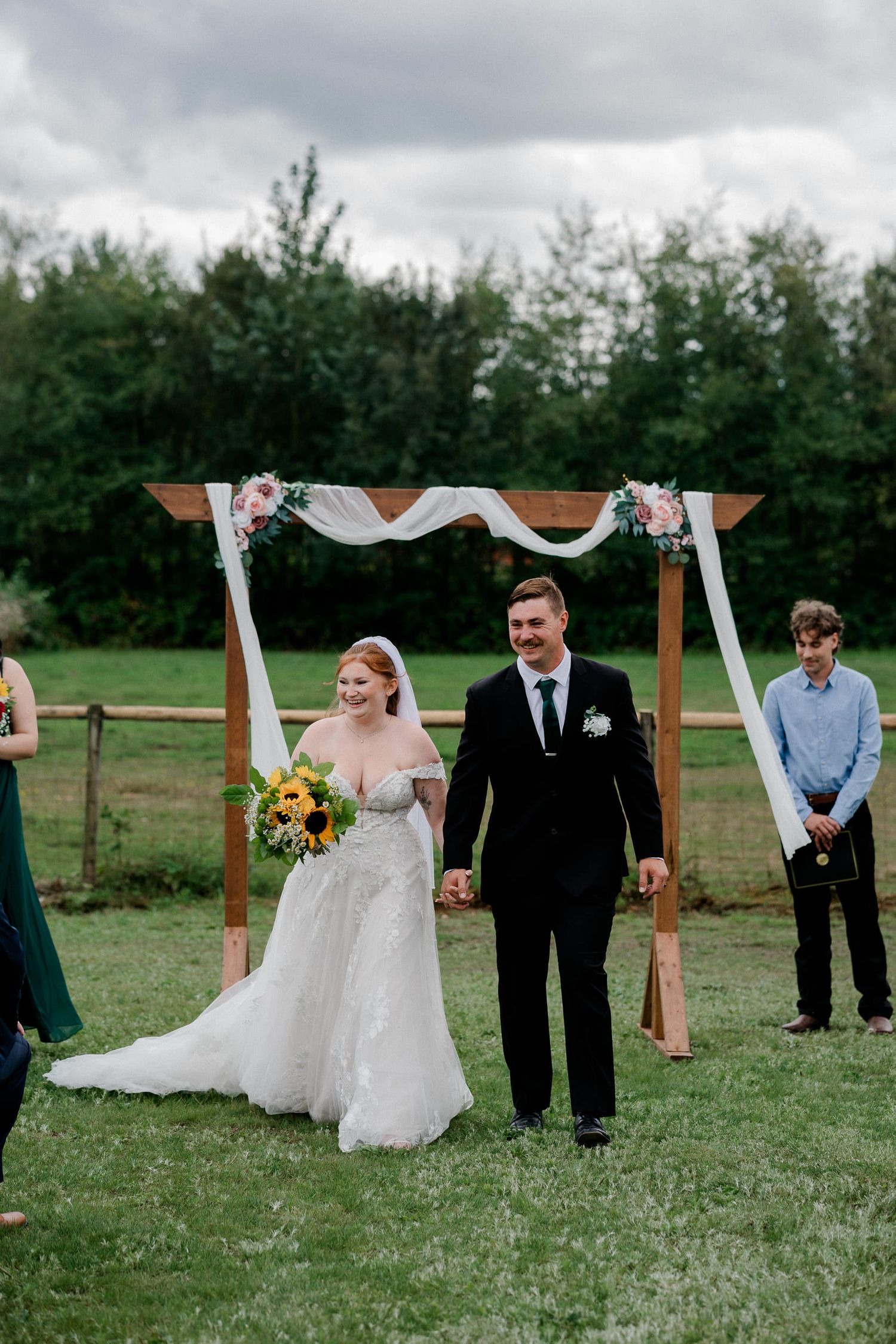 Wedding party stands outdoors under a rustic wooden arch decorated with white fabric and floral accents in a grassy field.
