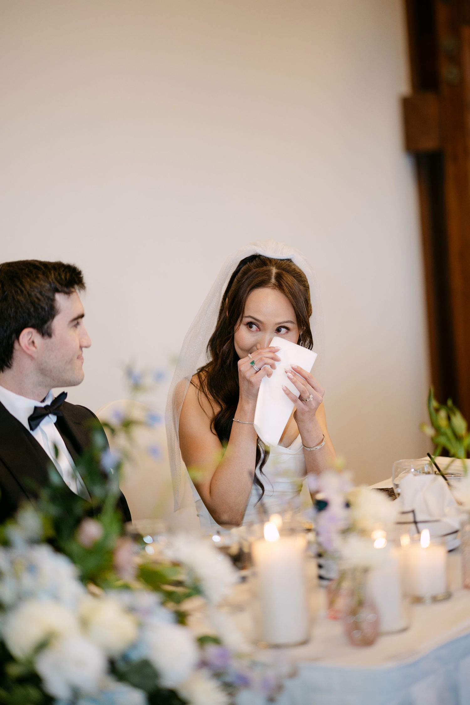 Emotional bride wiping tears at wedding reception table with groom beside her, surrounded by candles and floral arrangements.