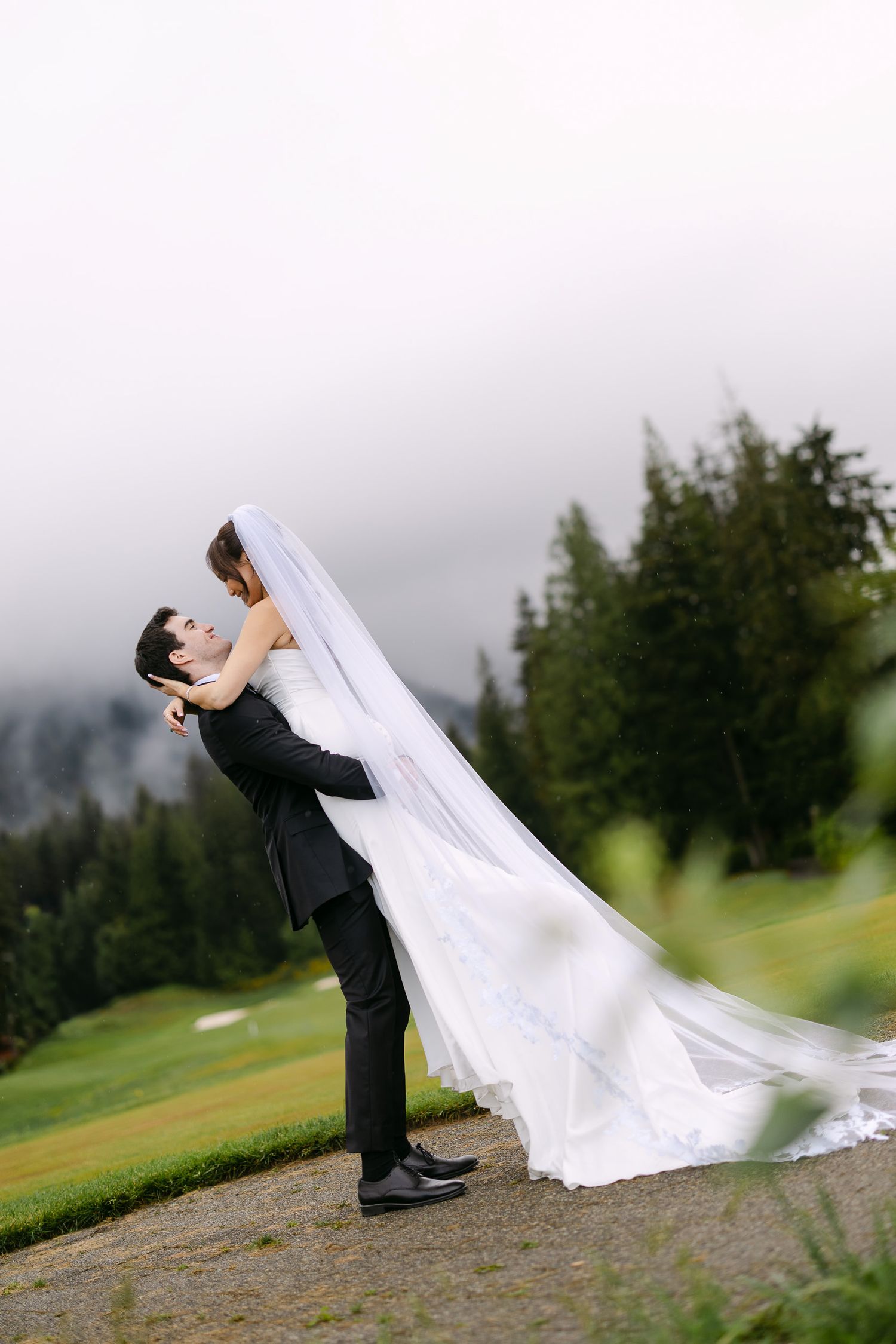 Groom lifts bride in white gown and veil outdoors with misty mountain trees backdrop.