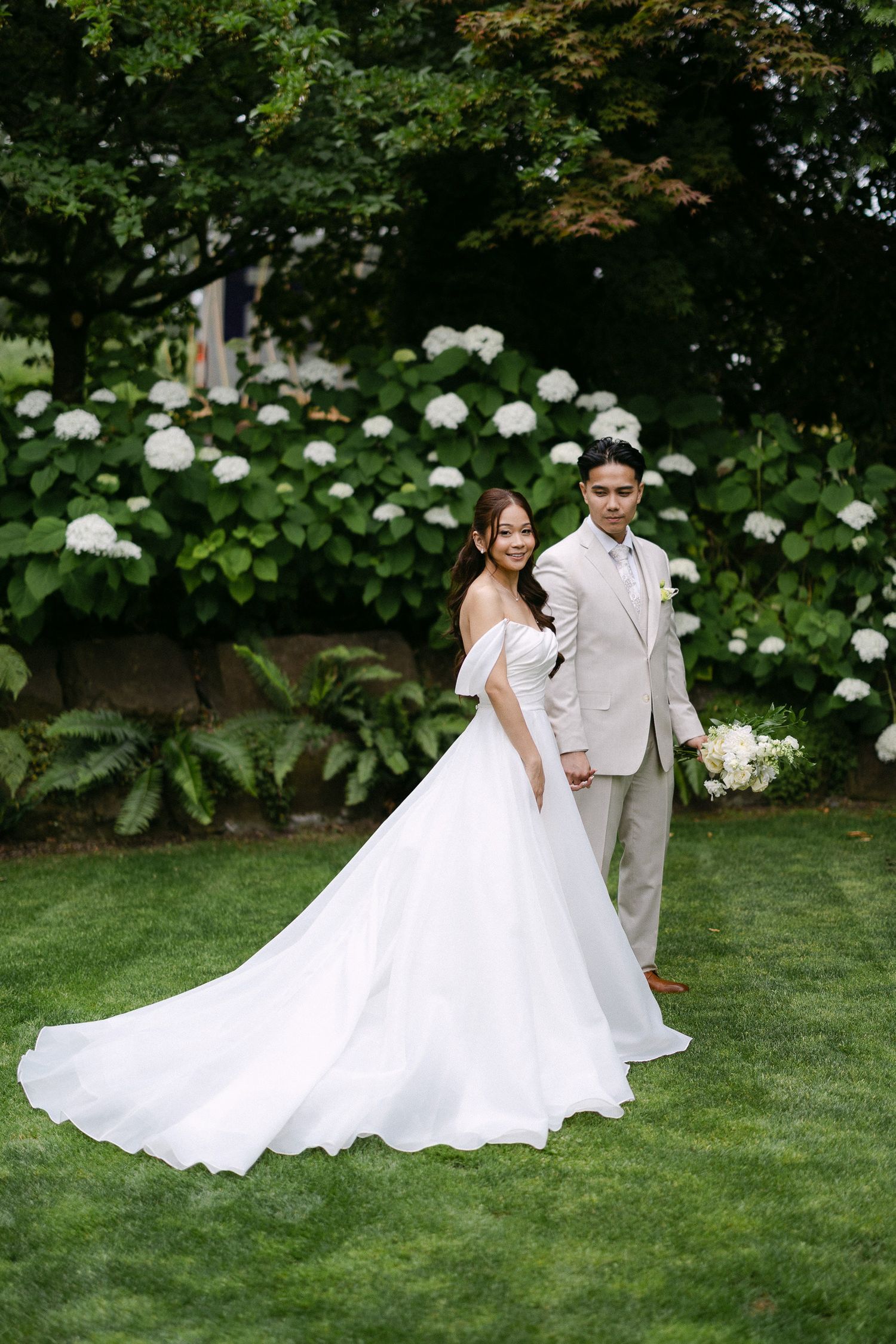 Bride in flowing white off-shoulder gown with long train poses with groom in beige suit before white hydrangeas.