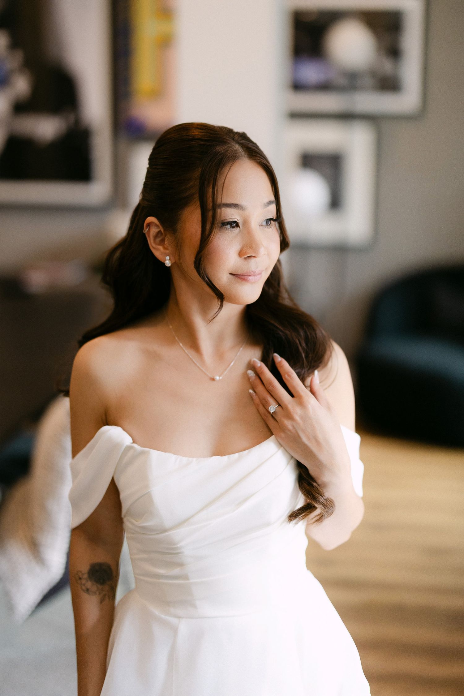 Bride in off-shoulder white gown with tattoo, wearing diamond ring and necklace, posing elegantly indoors.