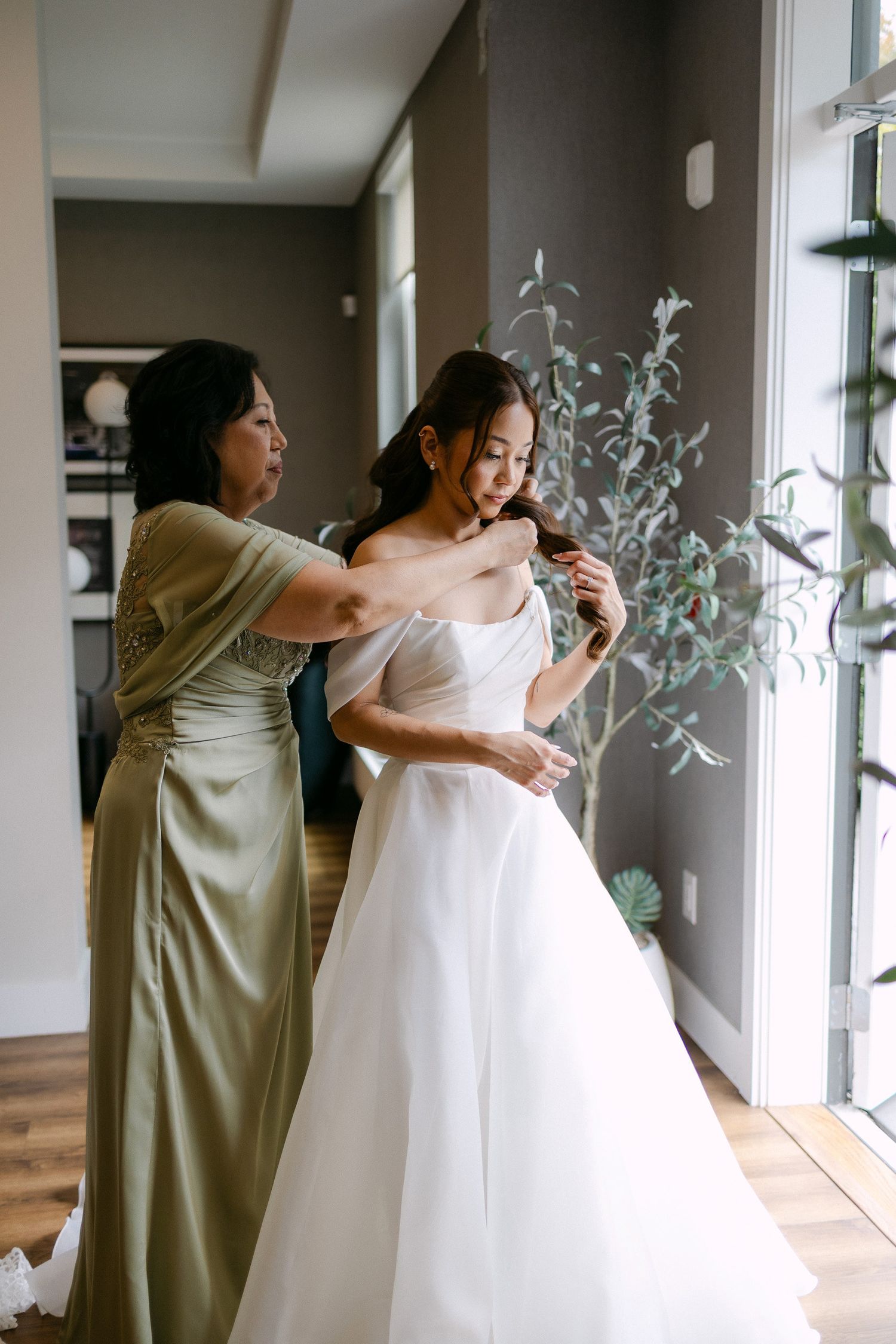 Mother helping bride into elegant strapless white ball gown wedding dress during bridal preparation at home.