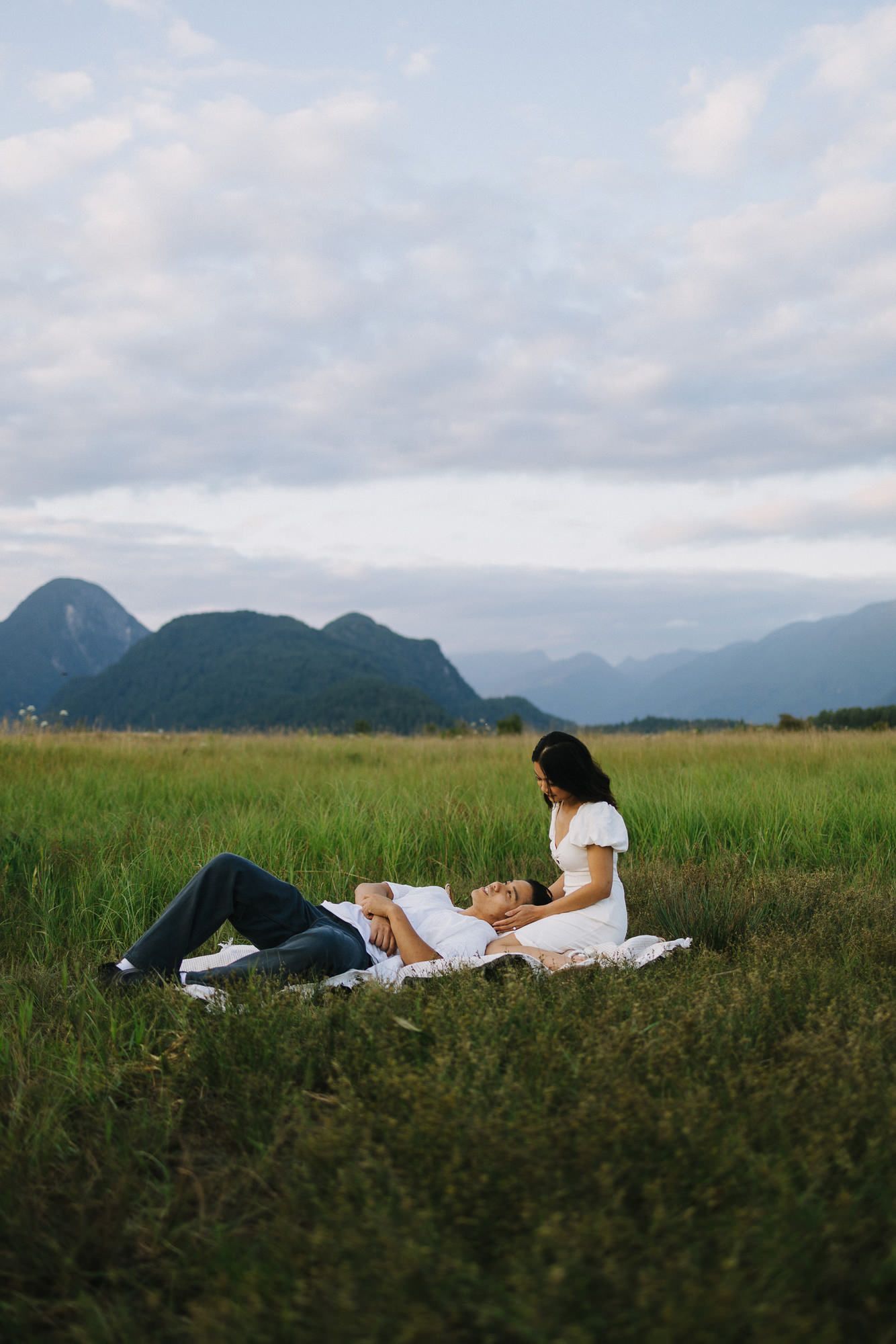 A couple in white lies together on a grassy field beneath a cloudy sky with mountains in the distance.