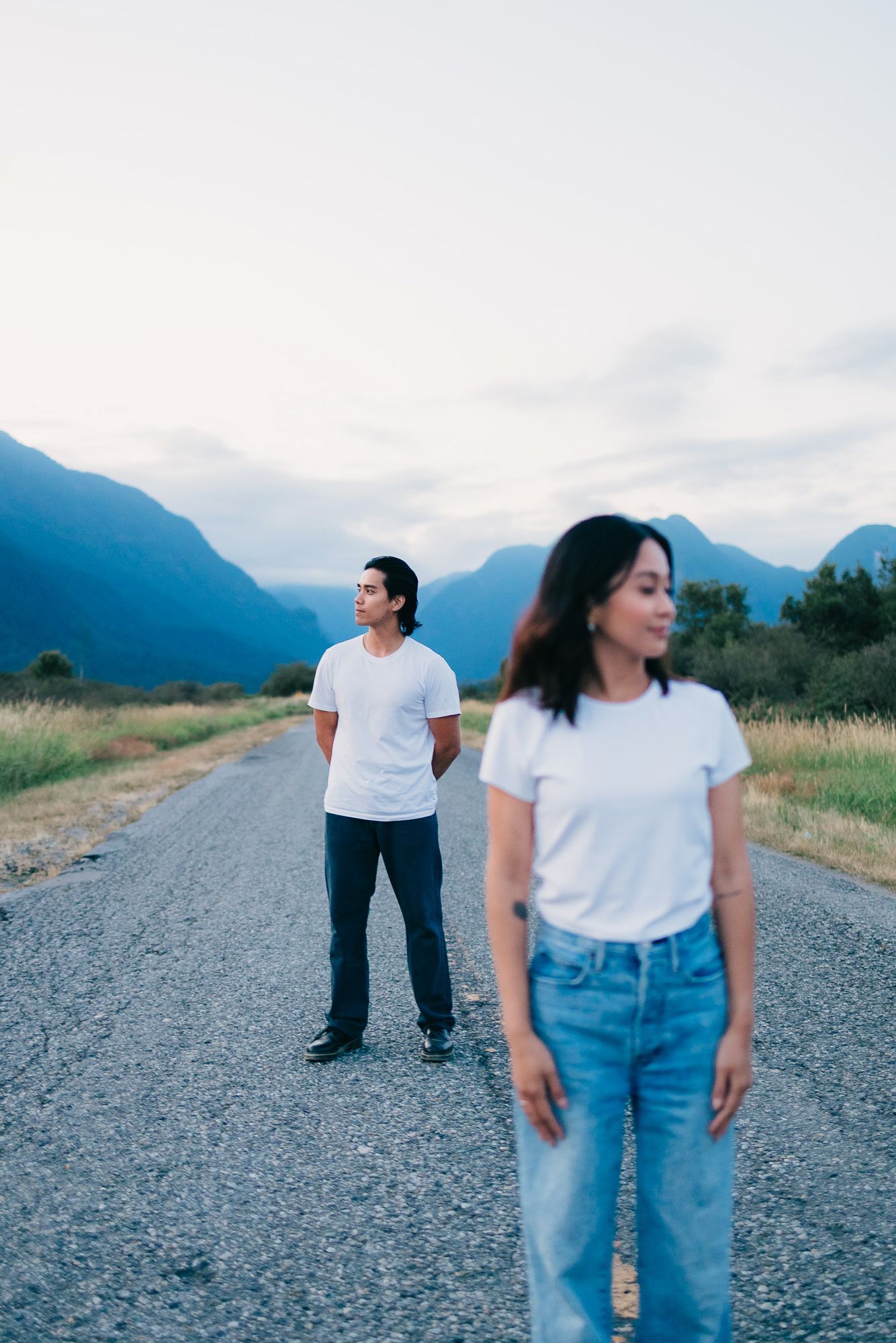 Woman in jeans stands in the foreground as a man looks away on a gravel road surrounded by blue mountains.