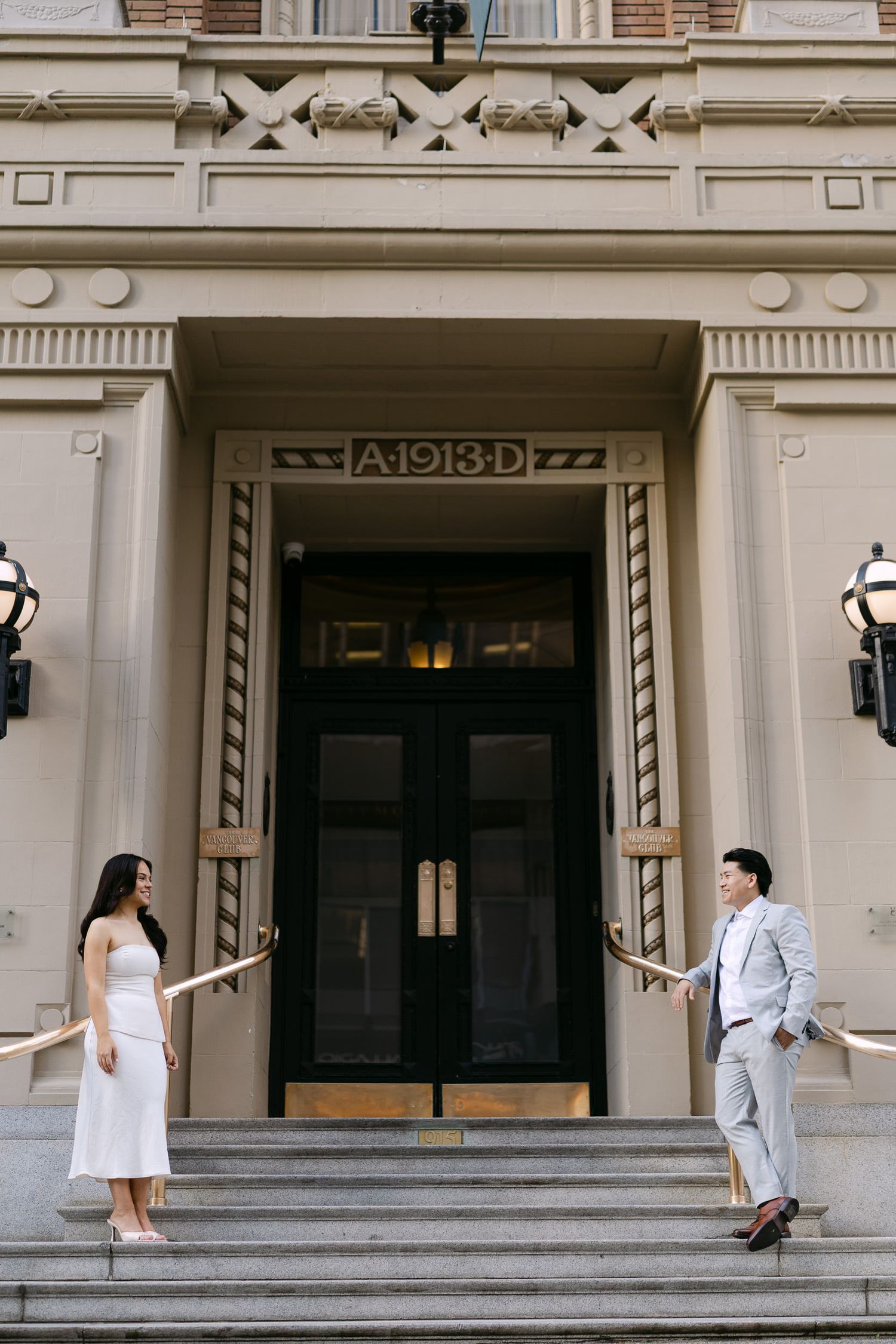 Engagement photo of a couple in a white dress and light gray suit posing on stone steps before a historic building entrance dated 1913.