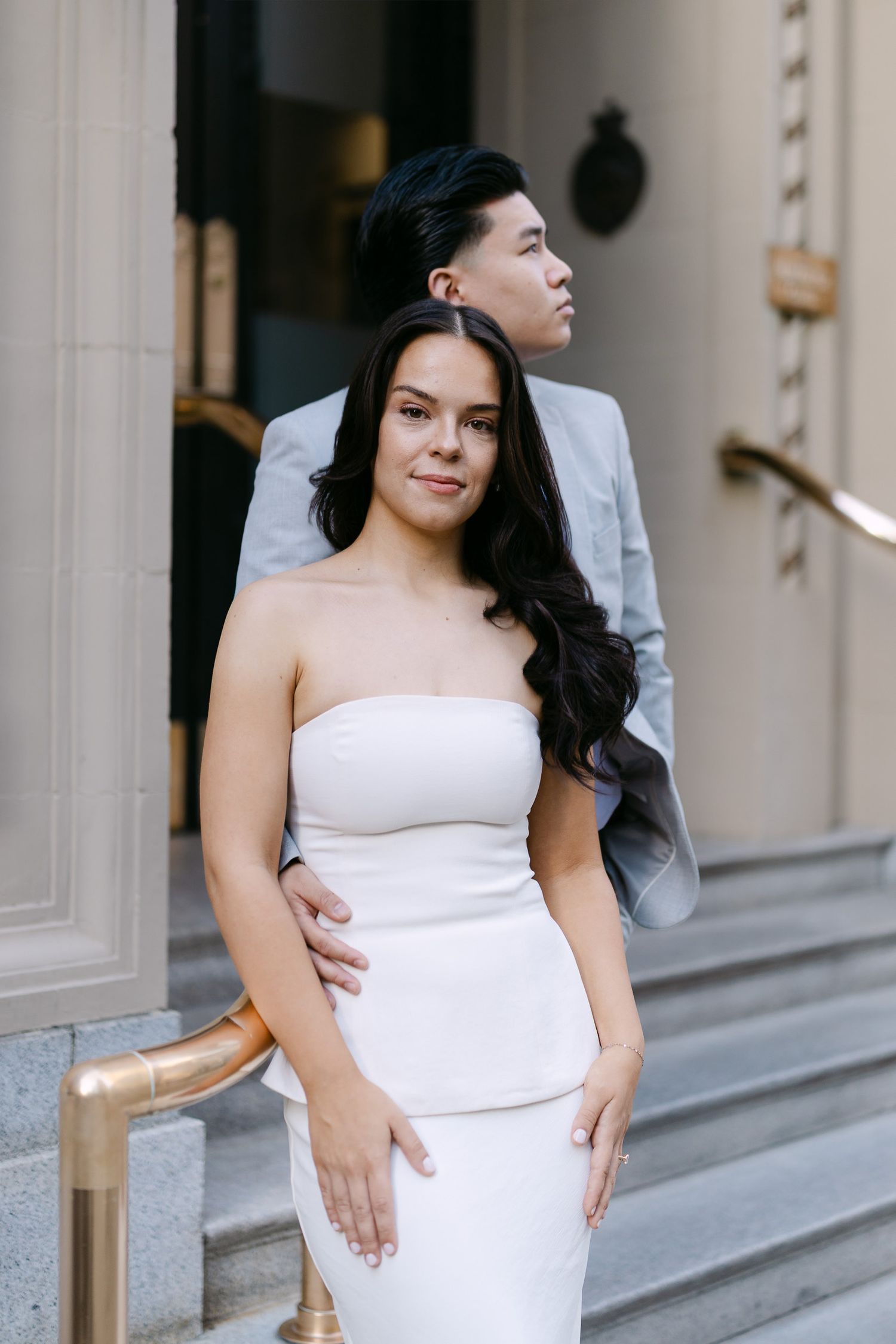 Couple posing on stone steps with a brass handrail, a woman in a white strapless dress and a man in a light gray suit behind her.