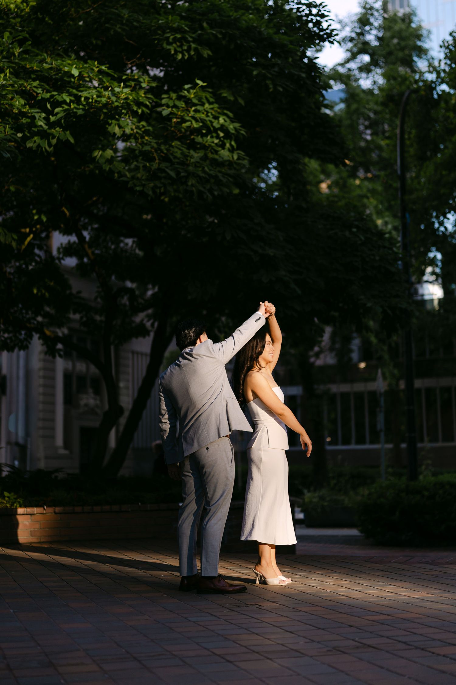 Man in a gray suit twirls a woman in a white dress on a brick plaza surrounded by trees in golden light.
