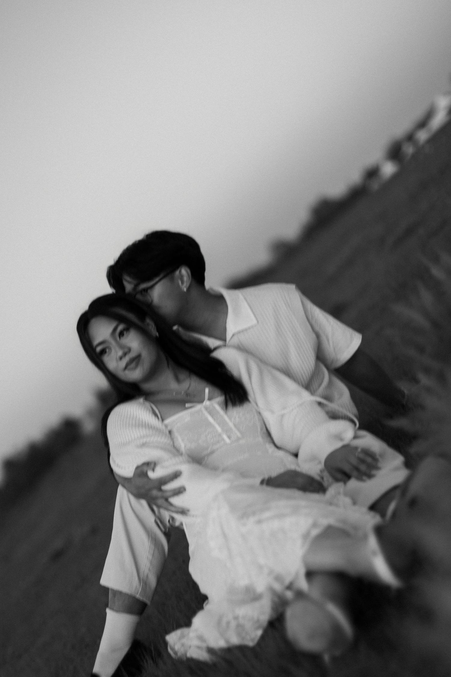 Black and white photo of a couple sitting close on a grassy hill, the man embracing a woman in a white lace dress at dusk.
