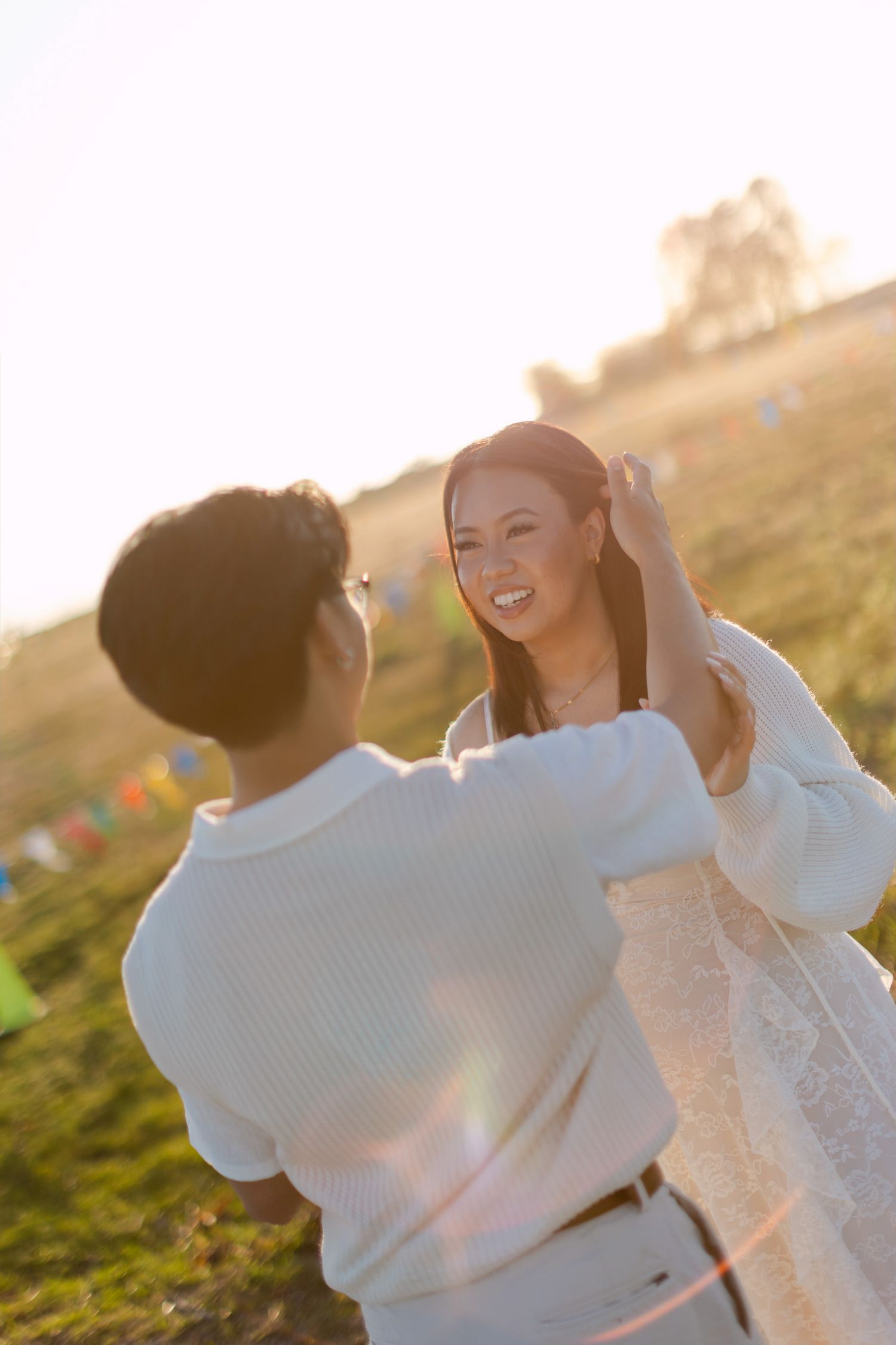 Smiling woman in a white lace dress laughs as a partner in a white shirt tenderly touches her hair in a sunlit grassy field at golden hour.
