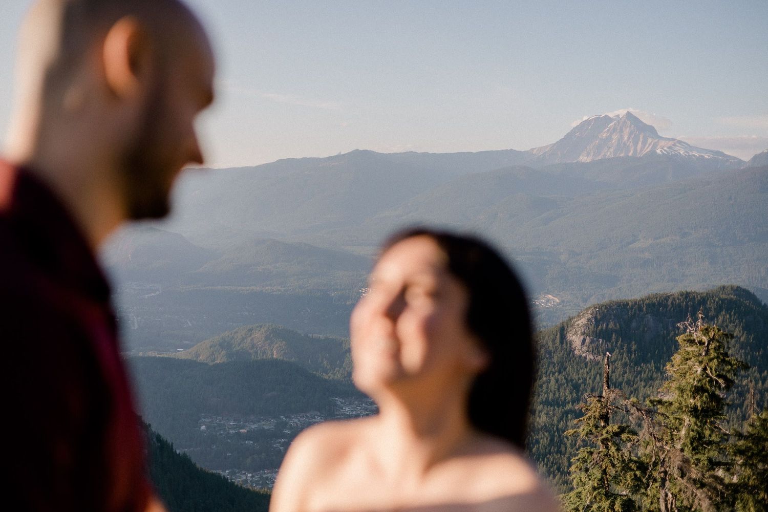 A couple shares a joyful moment on a mountain overlook with evergreen forests and distant peaks in the background.