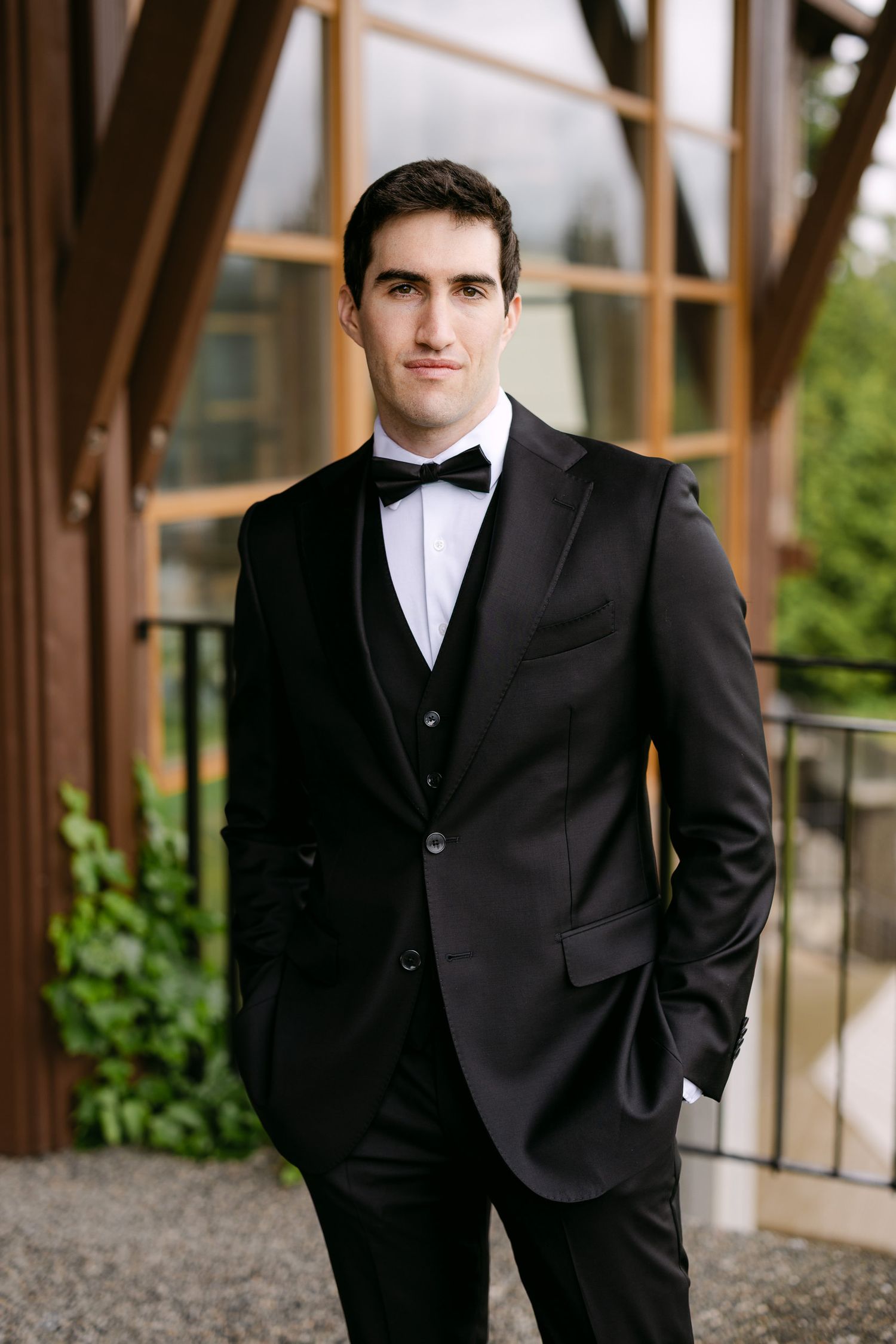 A groom in a black tuxedo with bow tie stands confidently outdoors near a modern wooden building.