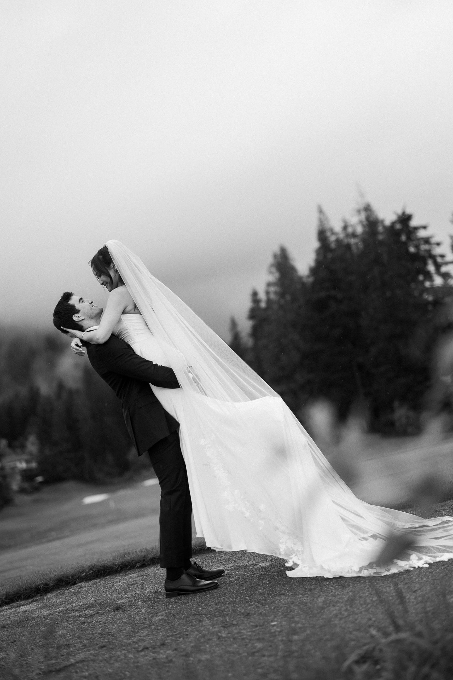 Black and white photo of groom lifting bride in flowing veil near evergreen trees.