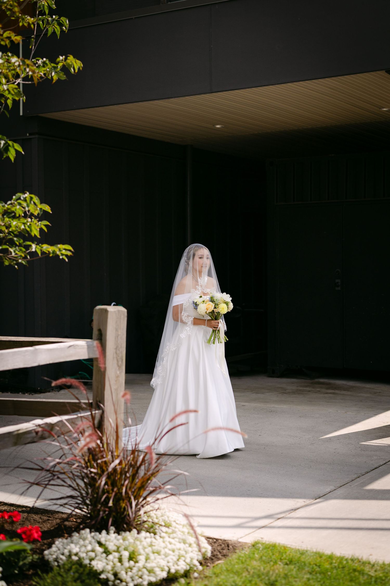 Bride in white off-shoulder gown and veil holds floral bouquet outside modern dark building with garden landscaping.