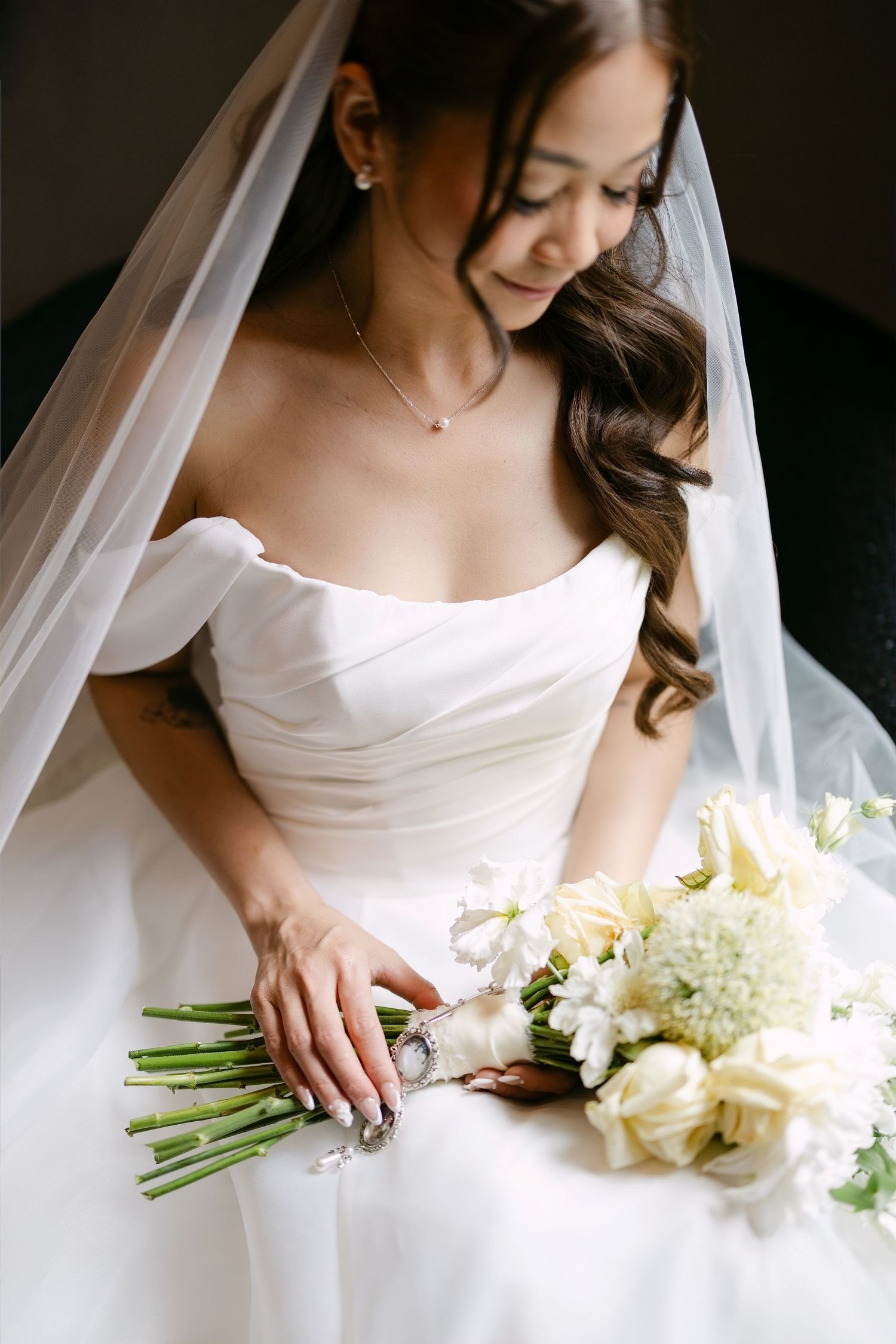 Bride in off-shoulder white gown and veil holds white floral bouquet, looking down with a serene smile.