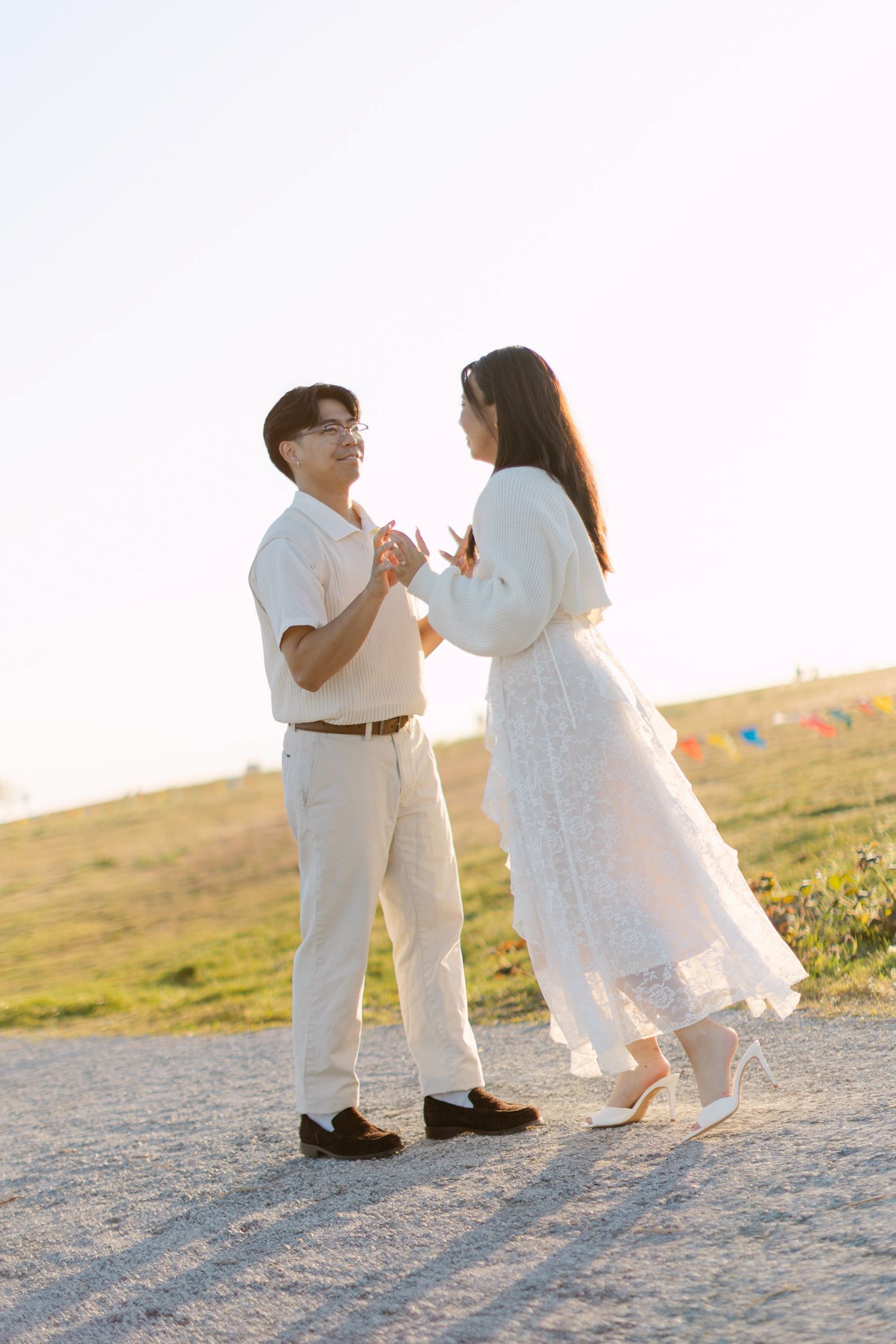 Couple in white and cream outfits holding hands and dancing on a gravel path with a grassy hillside and colorful bunting in the background.