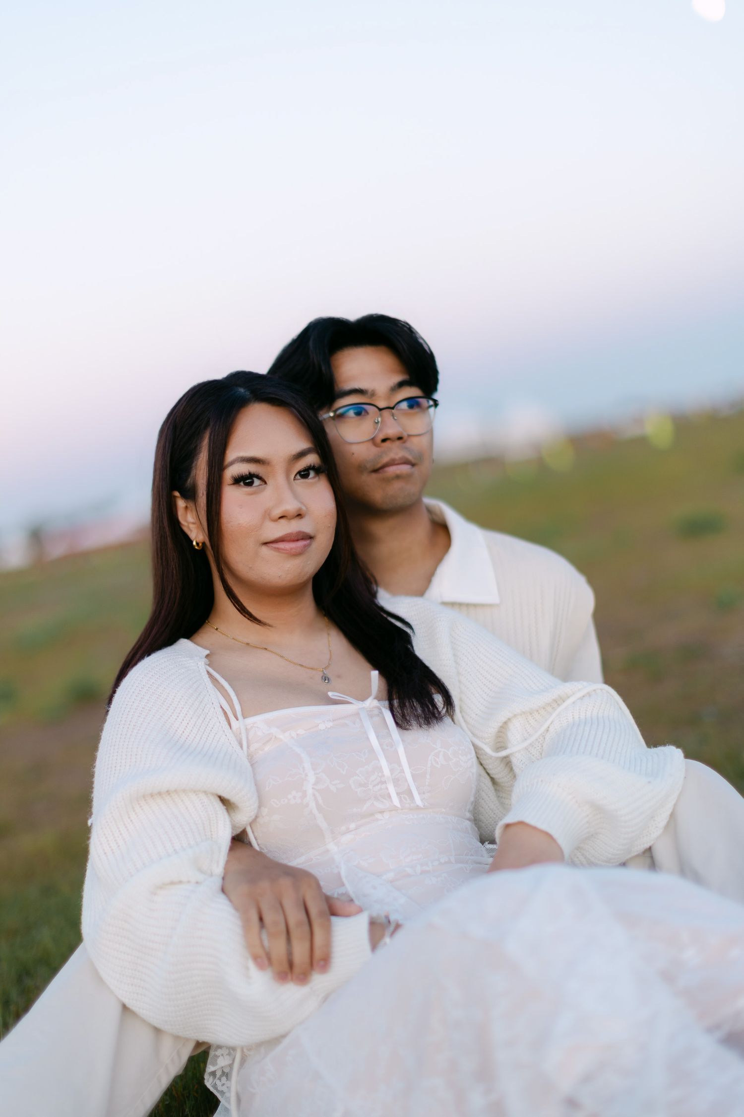 Couple in white outfits sitting close together on a grassy hillside at sunset, posing for a romantic engagement portrait.