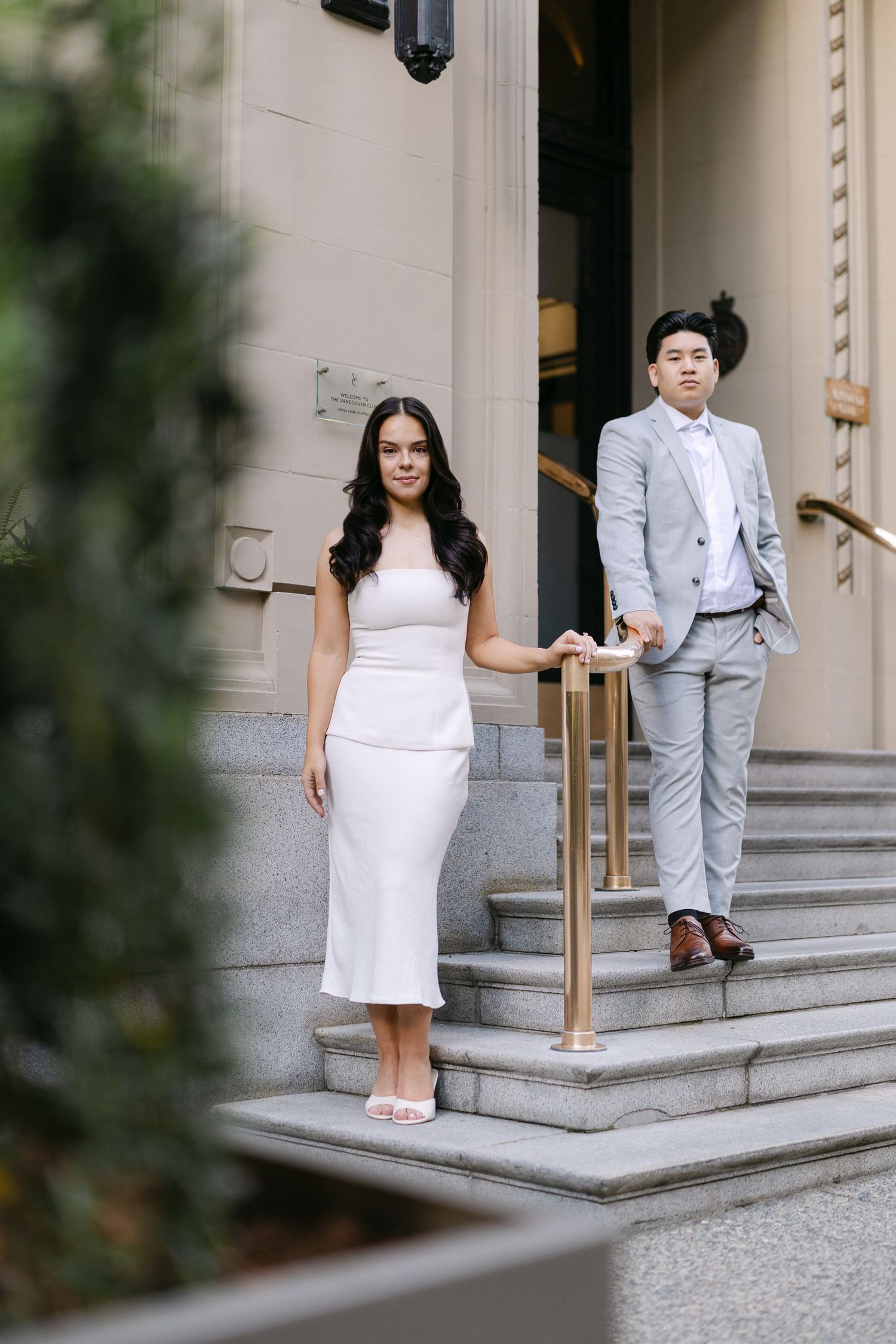 Woman in a white dress and man in a gray suit pose on stone steps with a brass handrail outside a historic building.