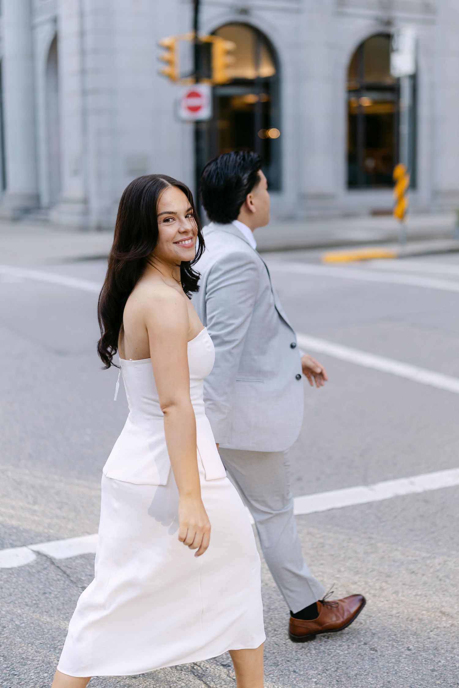 Smiling woman in a white dress glances back while walking across a city street beside her partner in a gray suit.