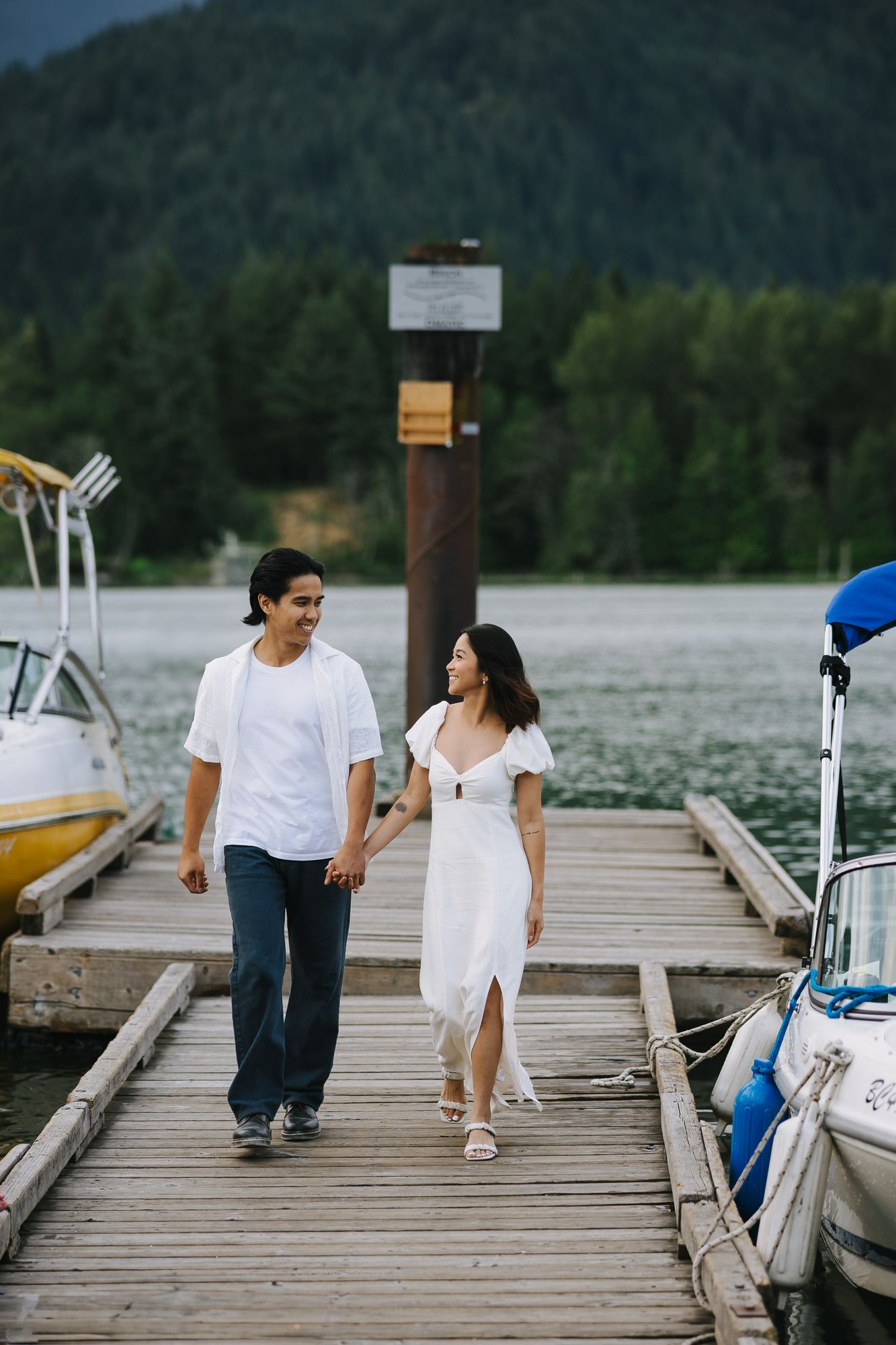 Smiling couple holding hands while walking along a wooden lakeside dock with boats and forested mountains in the background.