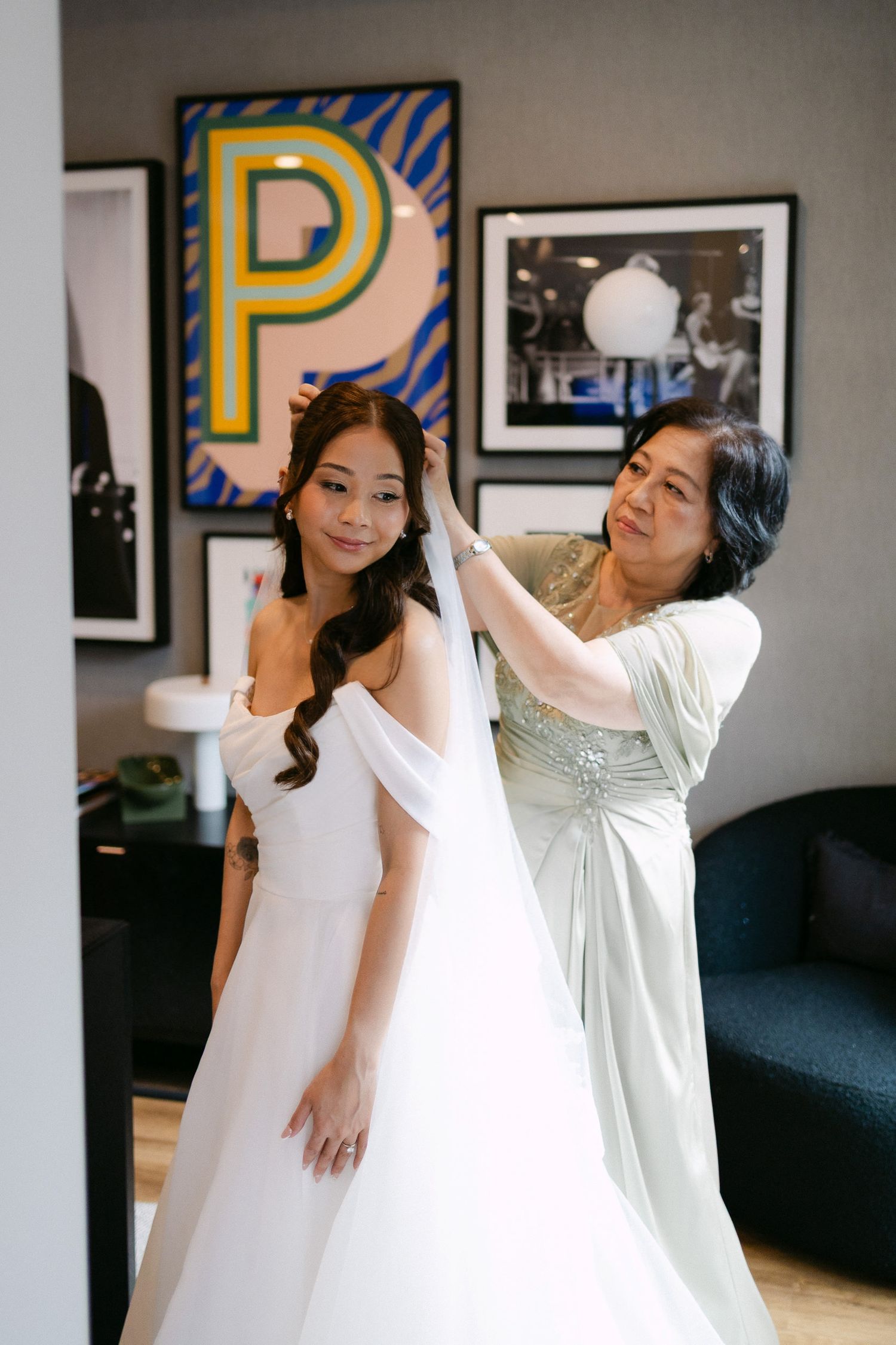 A bride in a white gown has her veil adjusted by an older woman in a sage dress in an elegant hotel room.