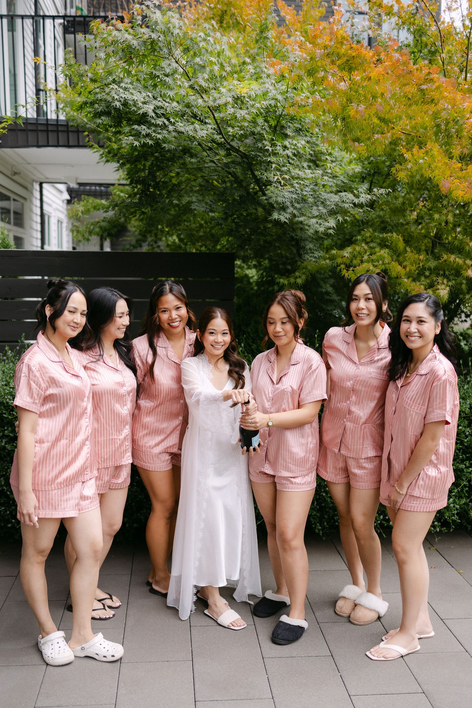 Bride and bridesmaids posing outdoors in matching pink satin pajama sets holding champagne.