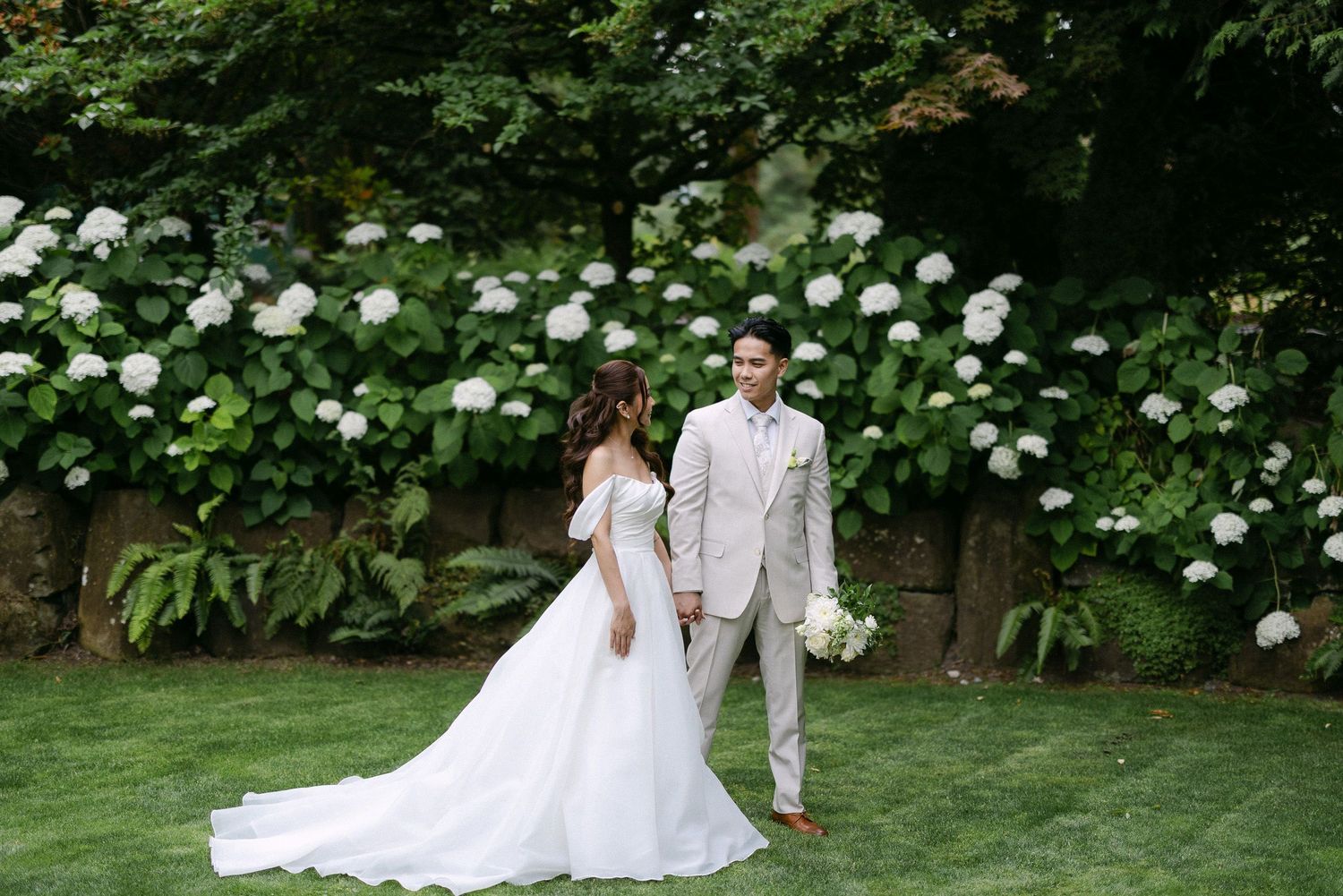 Bride and groom share a romantic first look moment on lush green lawn with white hydrangea bushes backdrop.