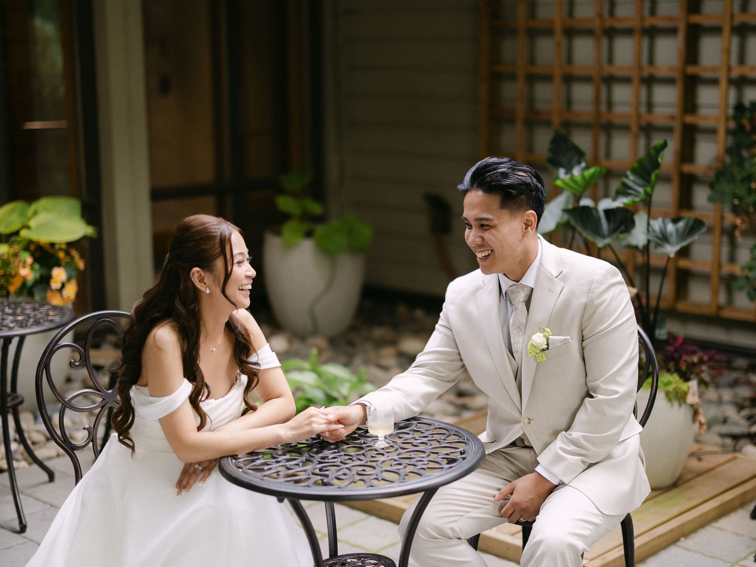 Smiling bride and groom in cream suit hold hands at ornate table in garden patio setting.