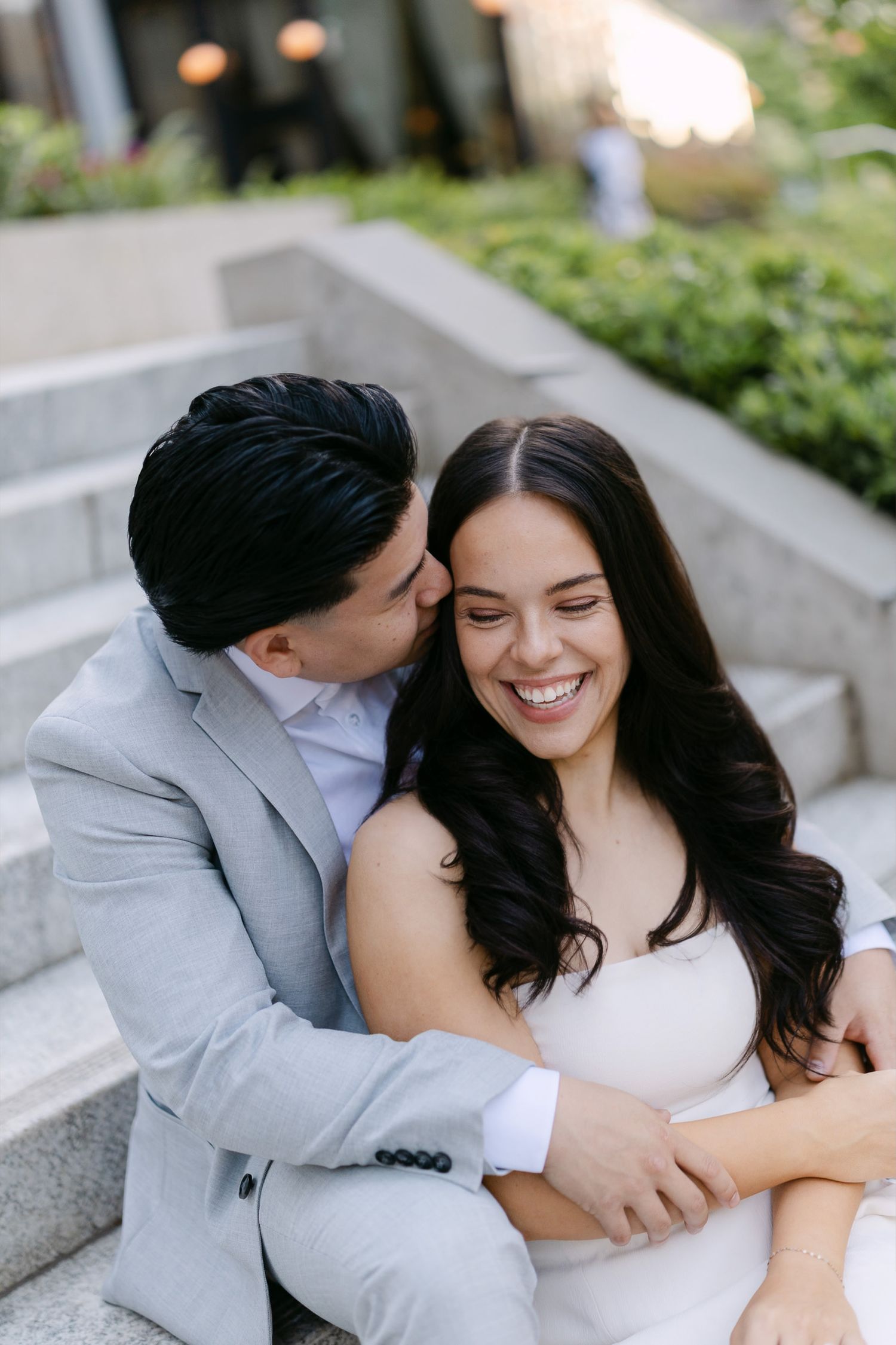 Smiling woman in a white dress embraced from behind by a man in a gray blazer as he kisses her cheek on outdoor stone steps.