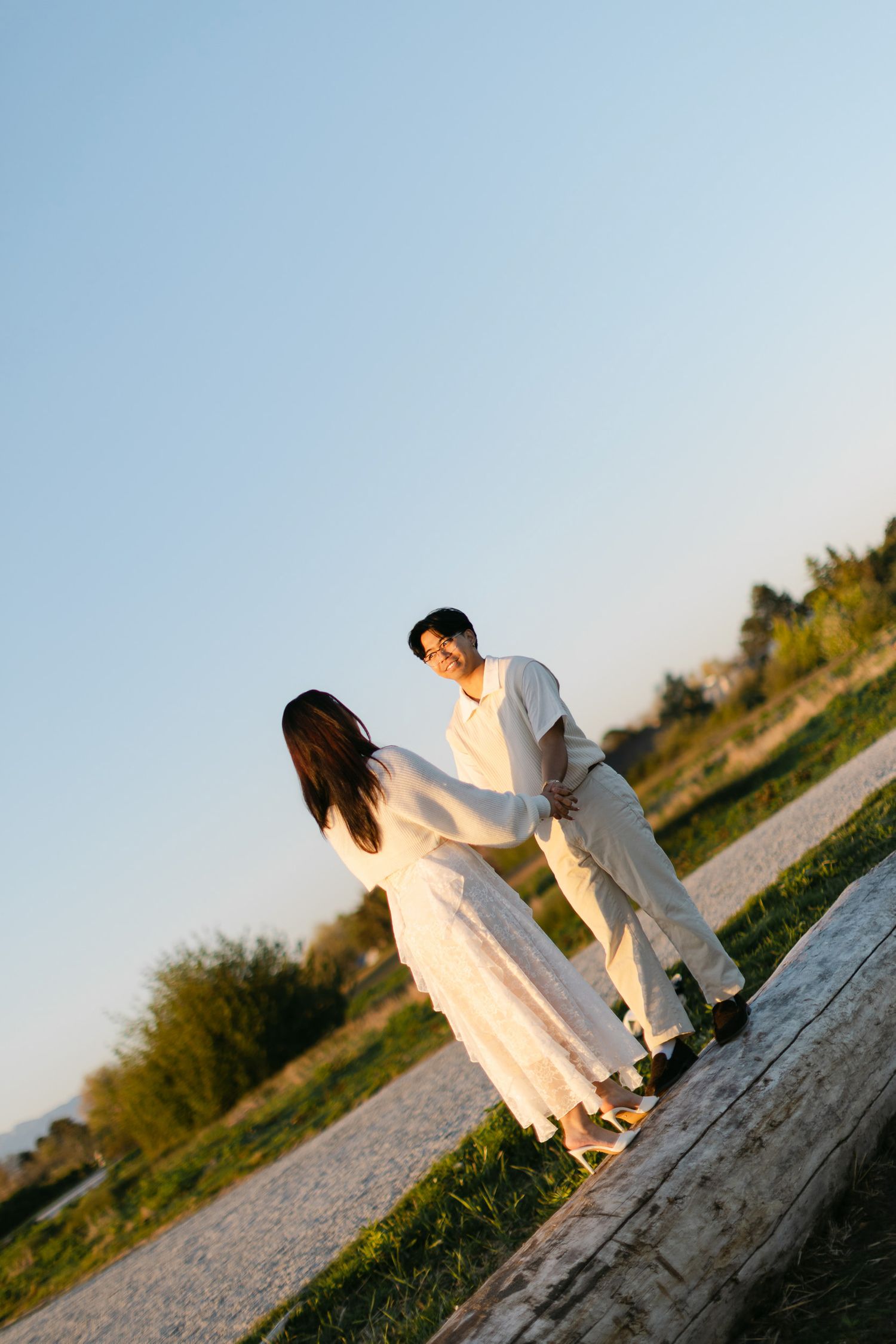 A couple in white outfits holds hands while walking along a gravel path beside a log, with grass and a clear evening sky behind them.