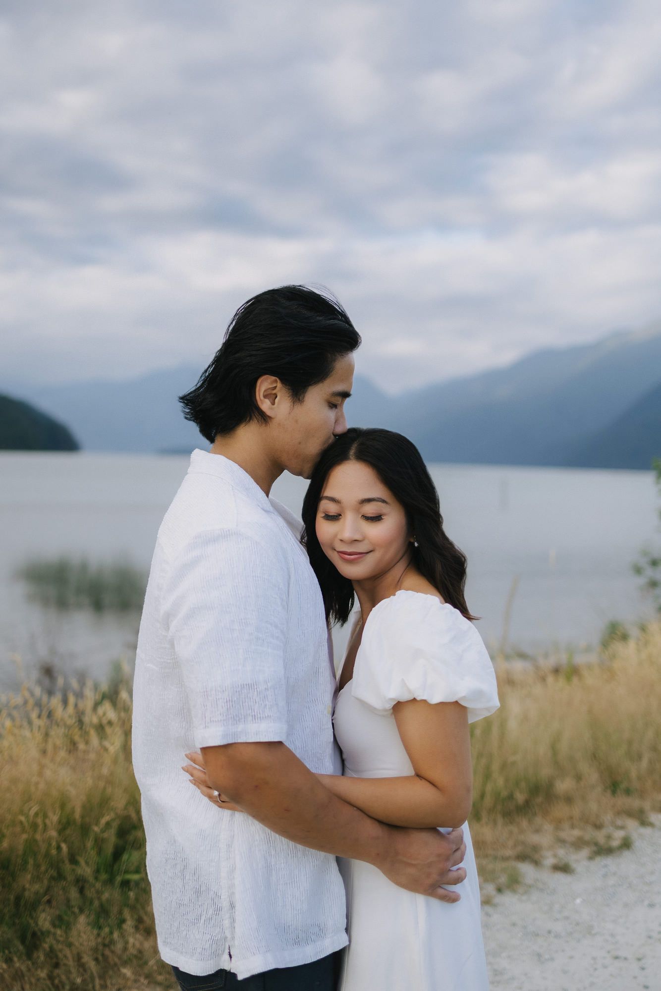 Man kisses his partner's forehead as they embrace beside a calm lake with misty mountains behind them.
