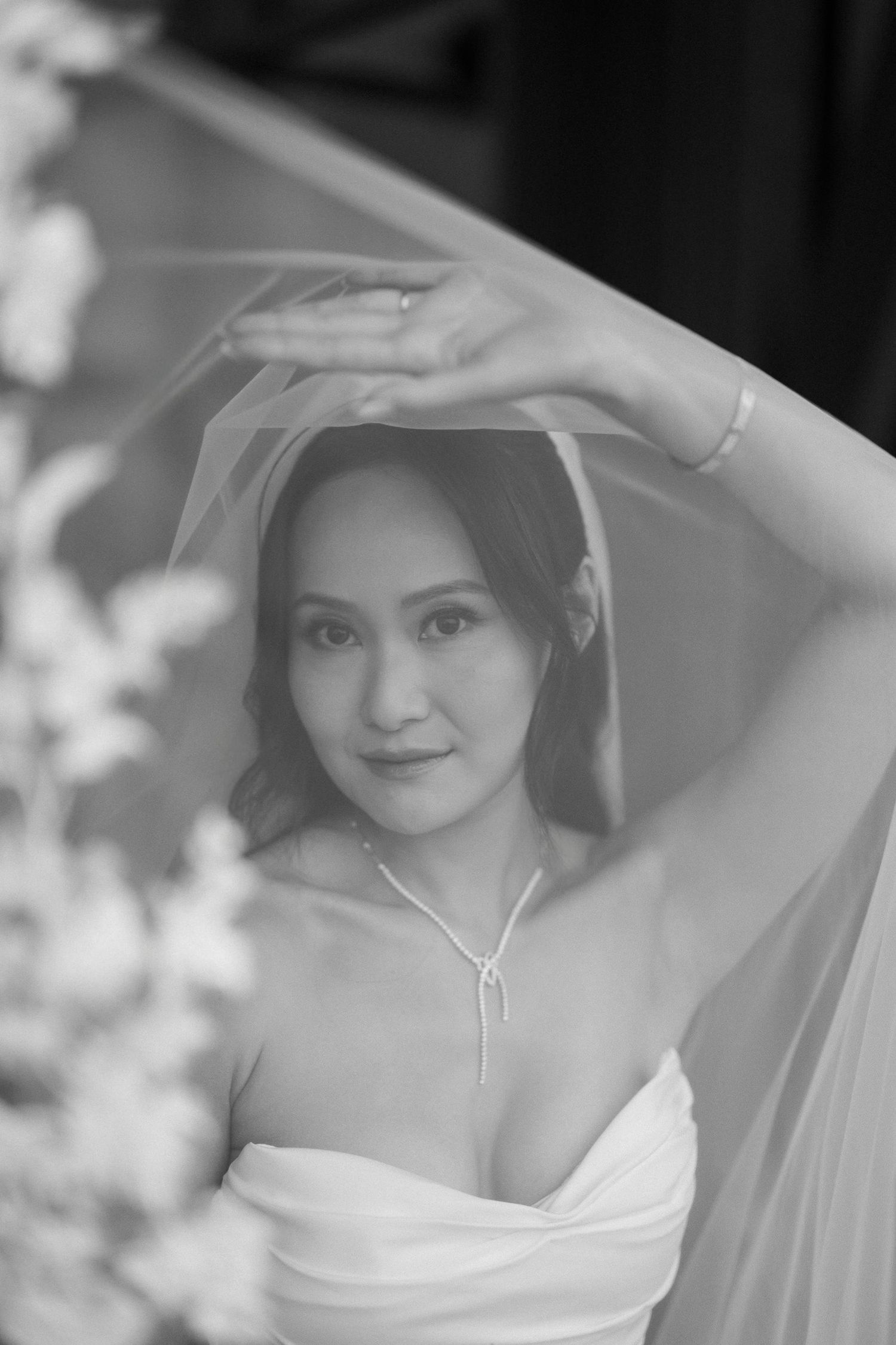 Black and white portrait of bride lifting veil with graceful arms in soft light.